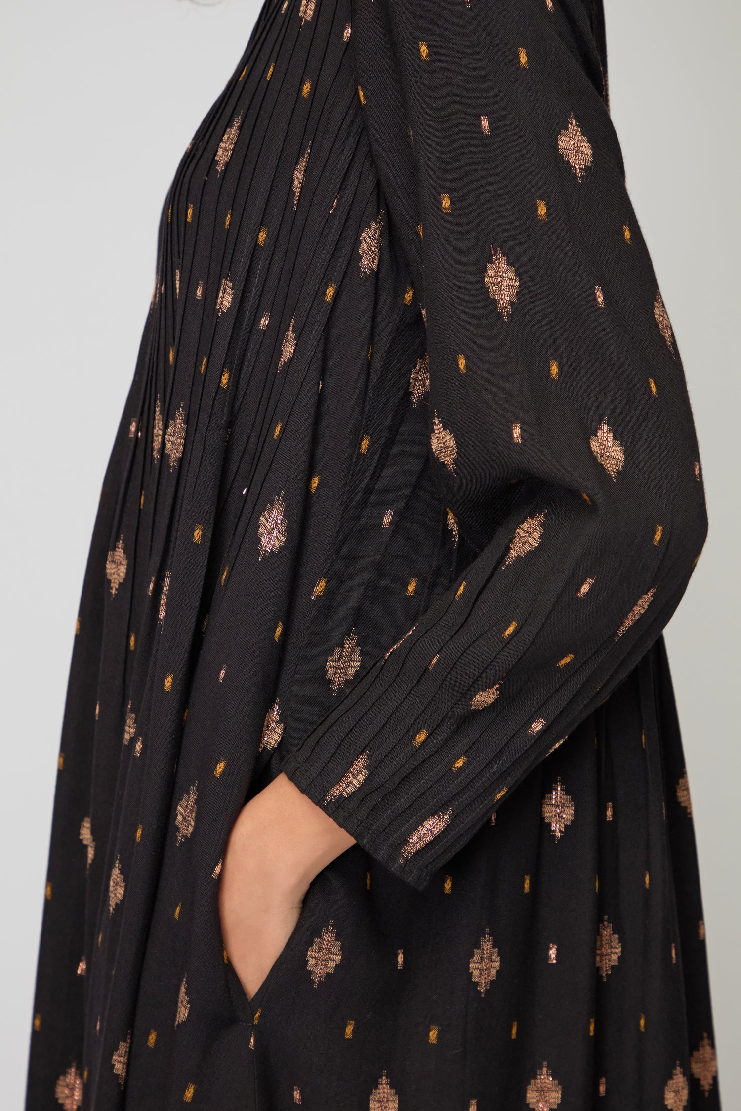 Someone wearing the Diamond Lurex Pleated Dress, featuring black and gold with bronze geometric patterns, places their hand into a side pocket.