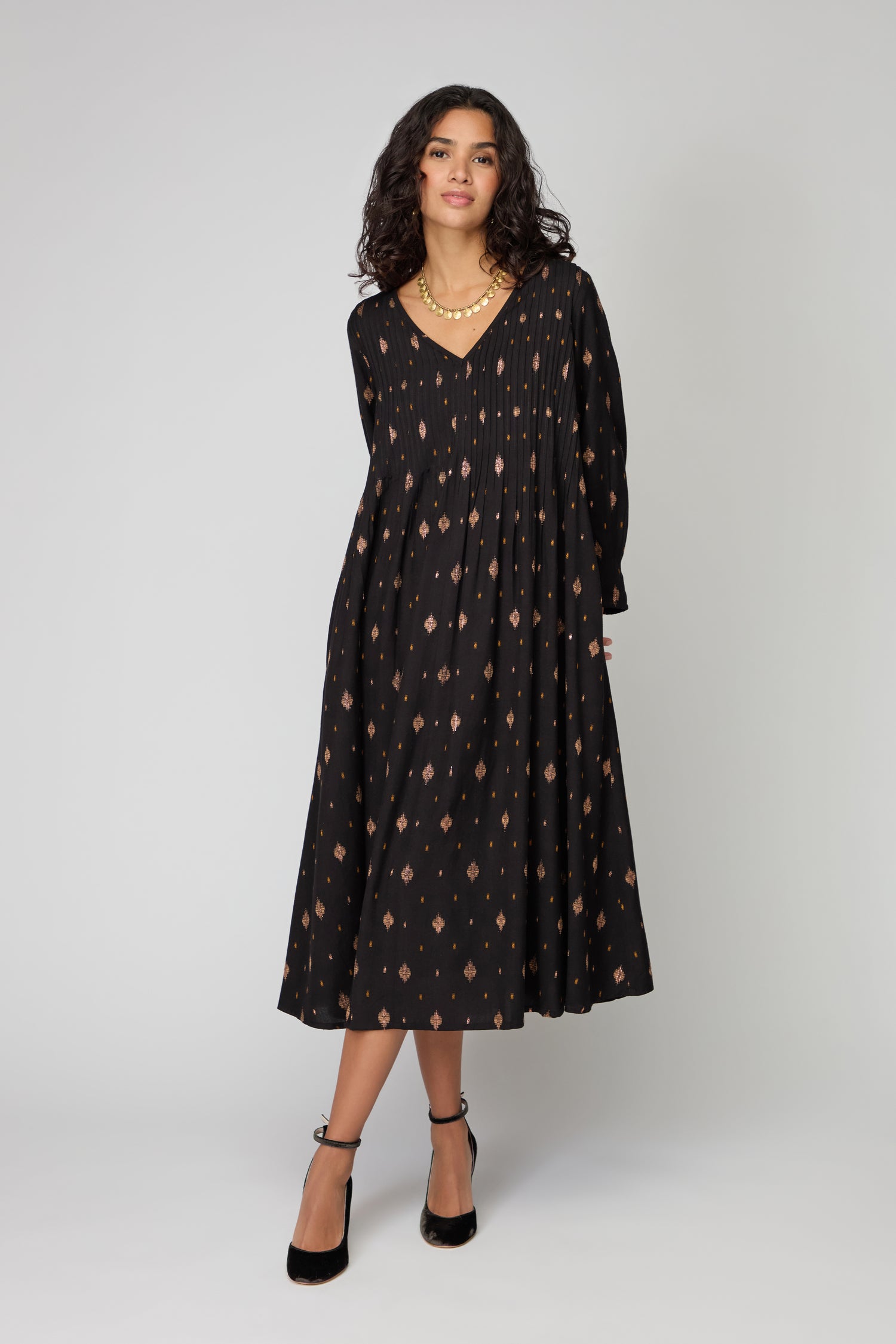 A woman models the Diamond Lurex Pleated Dress, featuring subtle geometric patterns in black and gold, styled with black ankle-strap heels and a gold necklace, against a plain light background.