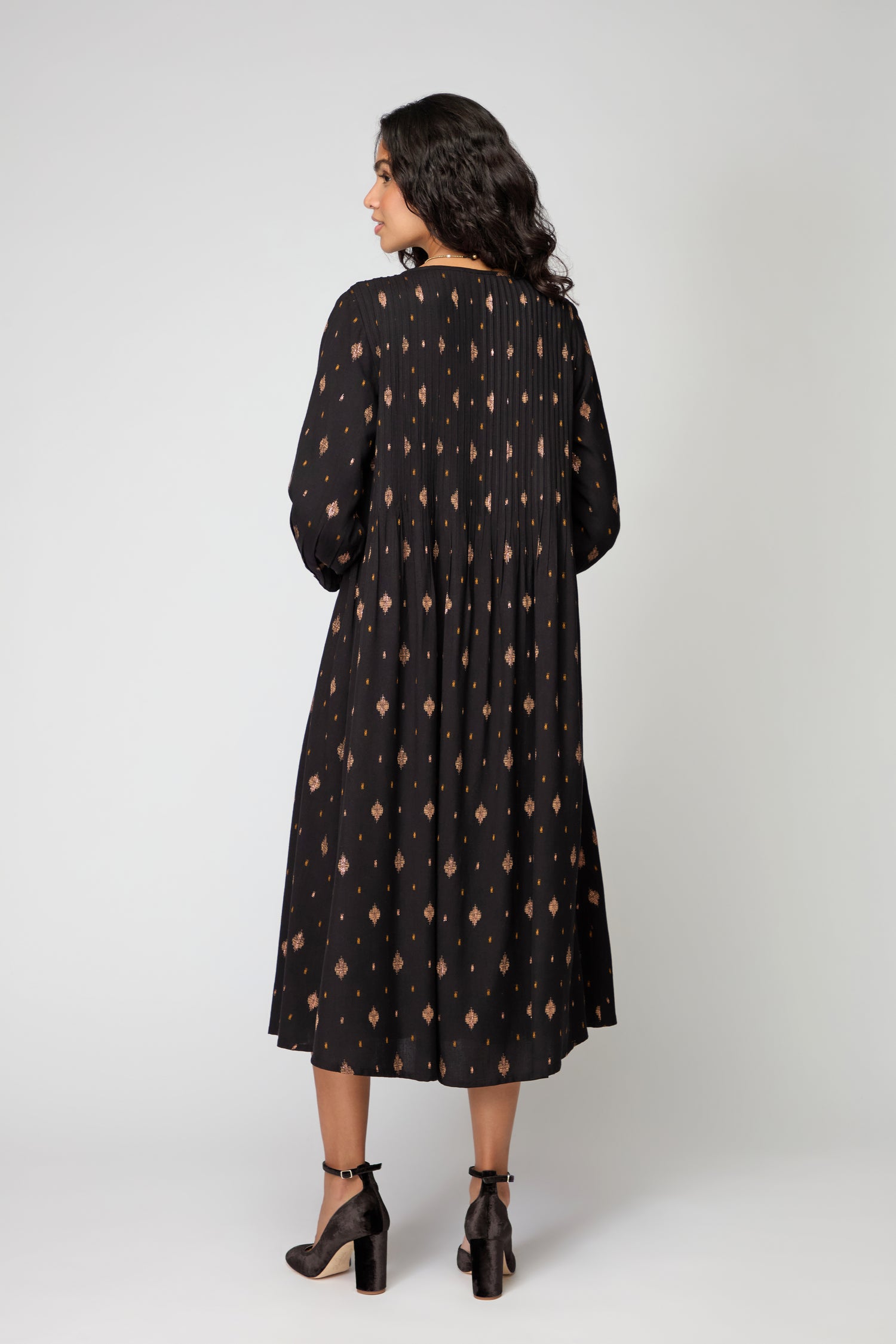 A woman stands in profile facing away, wearing the Diamond Lurex Pleated Dress—black and gold with a brown geometric pattern—and black heeled sandals against a plain light background.