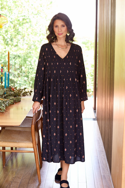 A woman in the Diamond Lurex Pleated Dress stands beside a wooden dining table in a bright room with large windows.