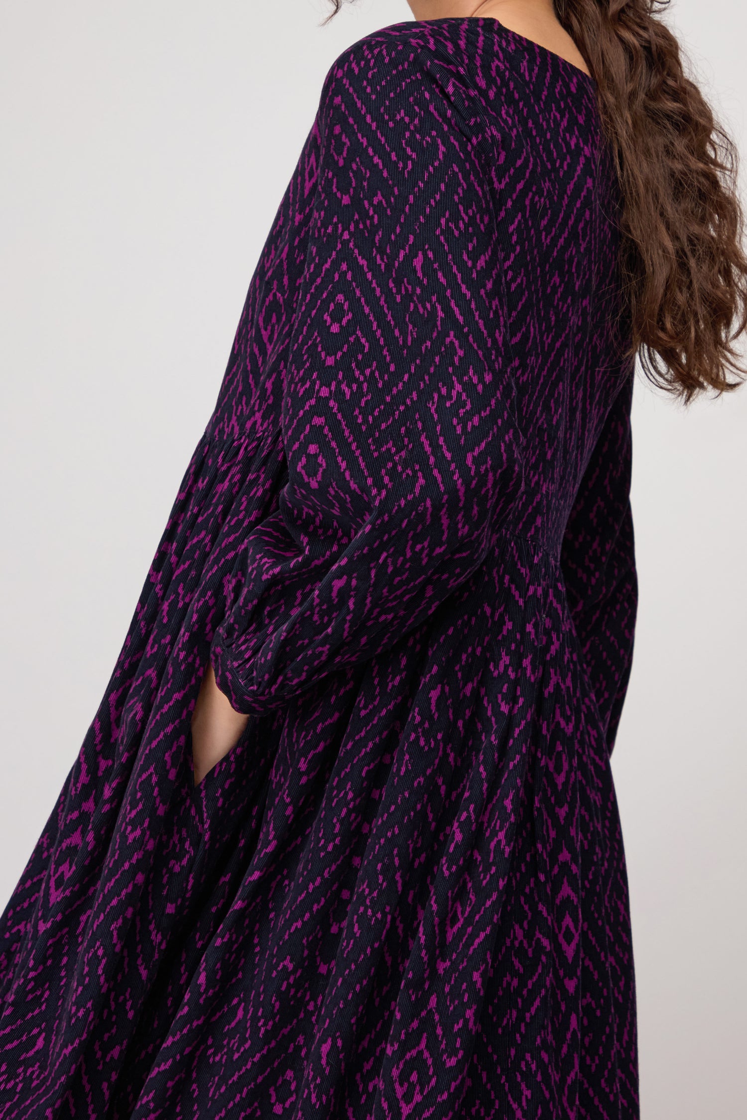 Model shown from the side wearing the Ornate Textured Plum Cord Dress, which features a relaxed silhouette and a geometric purple and black pattern, with one hand placed in the pocket.