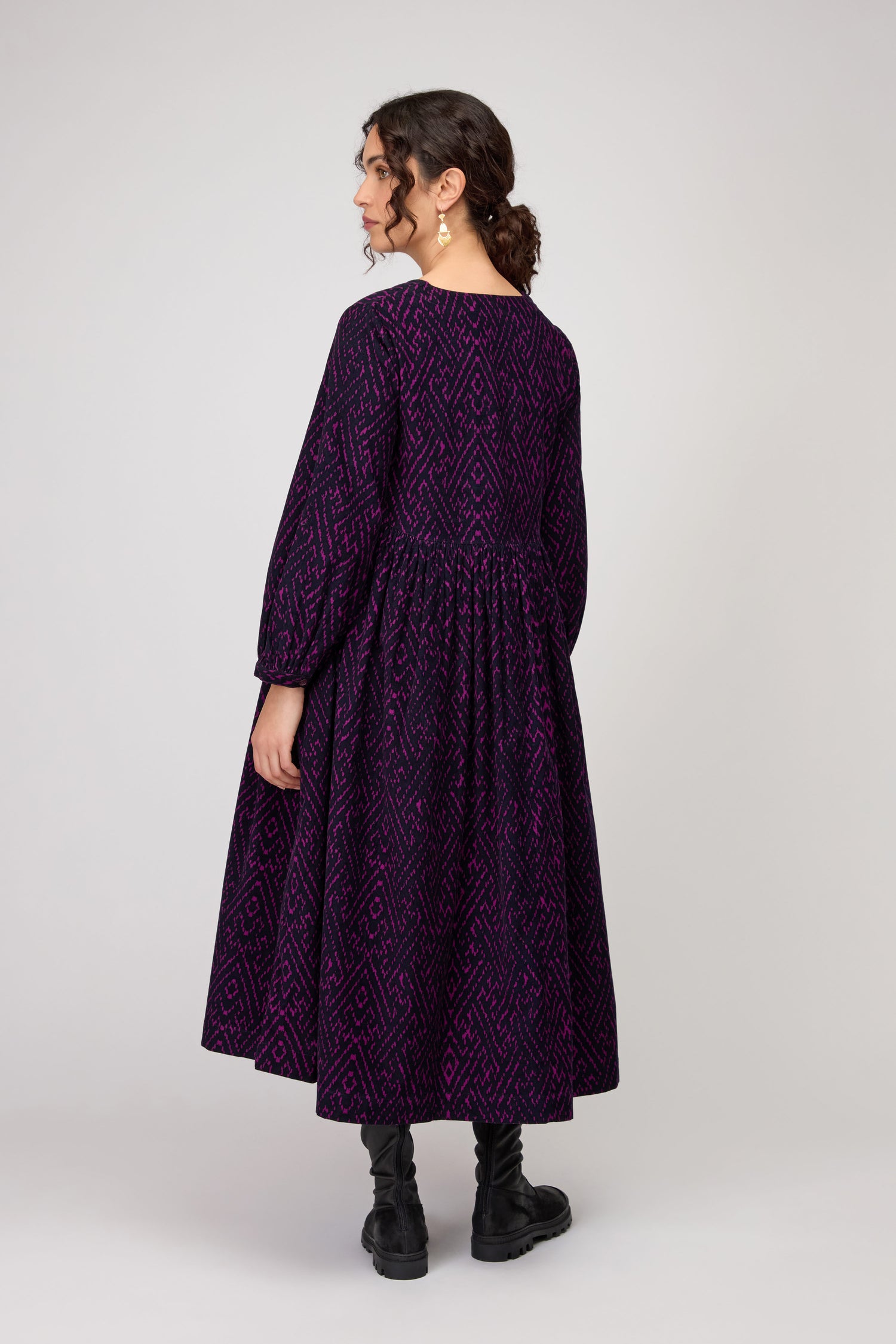 A woman faces away wearing the Ornate Textured Plum Cord Dress, featuring a relaxed silhouette, purple pattern, flared skirt, and paired with black boots—an effortlessly versatile choice against a plain light background.