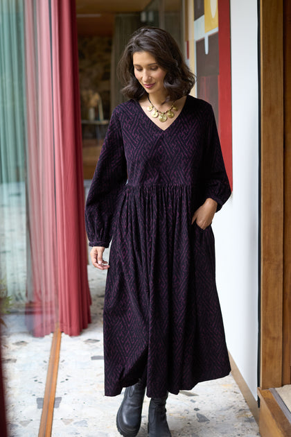A woman stands indoors wearing the Ornate Textured Night and Plum Cord Dress with a relaxed silhouette, styled with black boots and a gold necklace. She has one hand in her dress pocket and looks down.