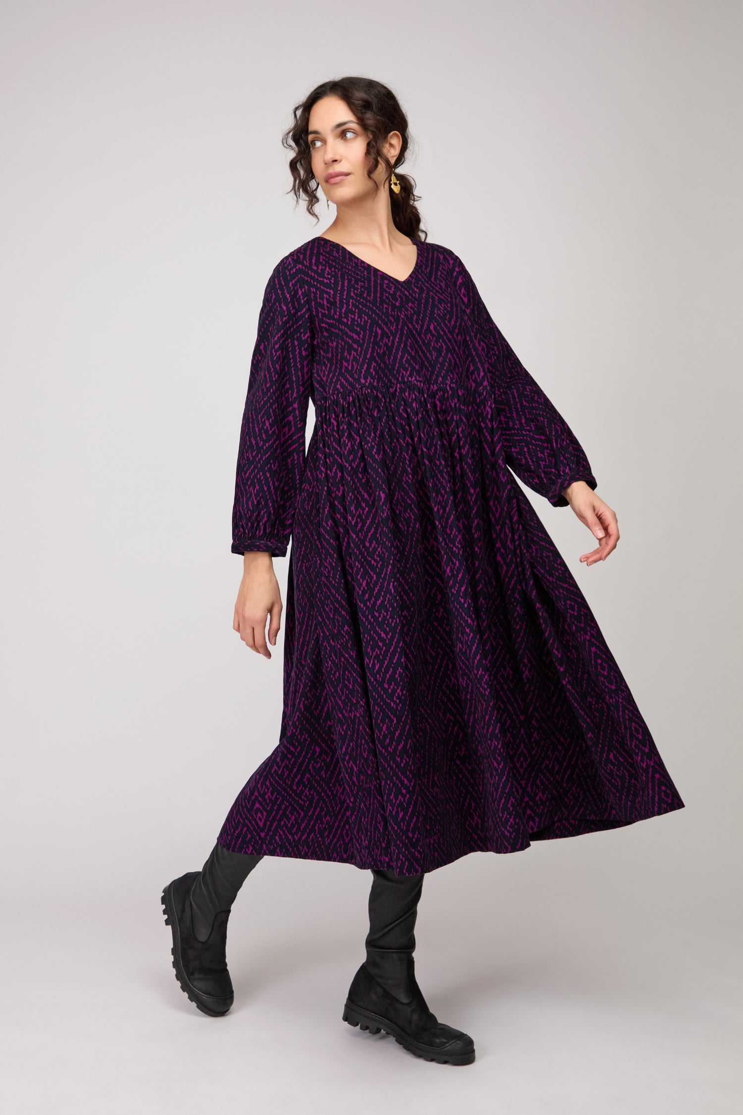 A woman stands against a plain background, looking left with one foot forward, wearing the Ornate Textured Plum Cord Dress—its long sleeves, purple pattern, and relaxed fit paired with black boots for a versatile look.