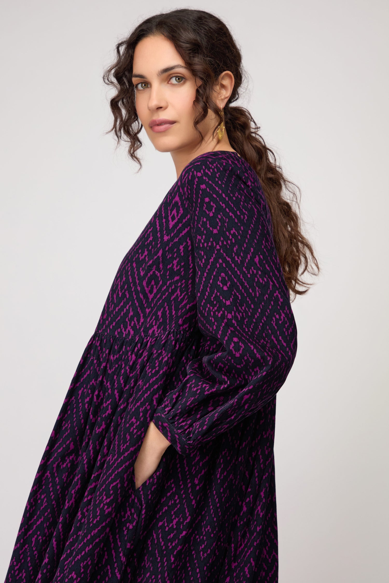 Woman with long curly brown hair wears the Ornate Textured Plum Cord Dress in purple and black, standing sideways with hands in pockets against a plain background, highlighting the dress’s relaxed silhouette.