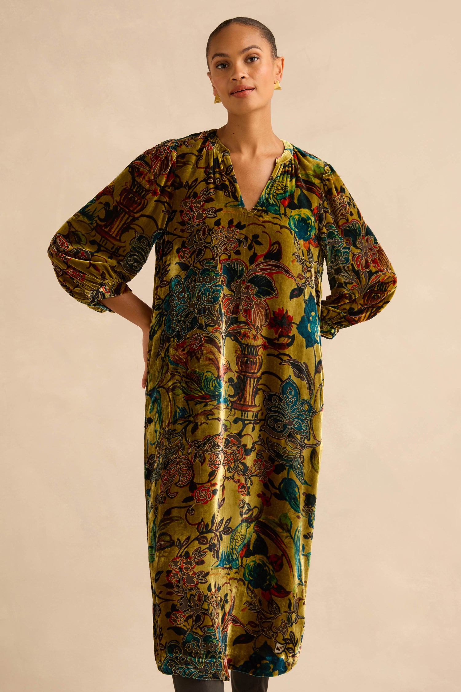 Model wearing a Silk Velvet Floral Devore Dress in olive green with a midi-length V-neck, three-quarter length sleeves, and a colorful floral and bird devoré pattern.
