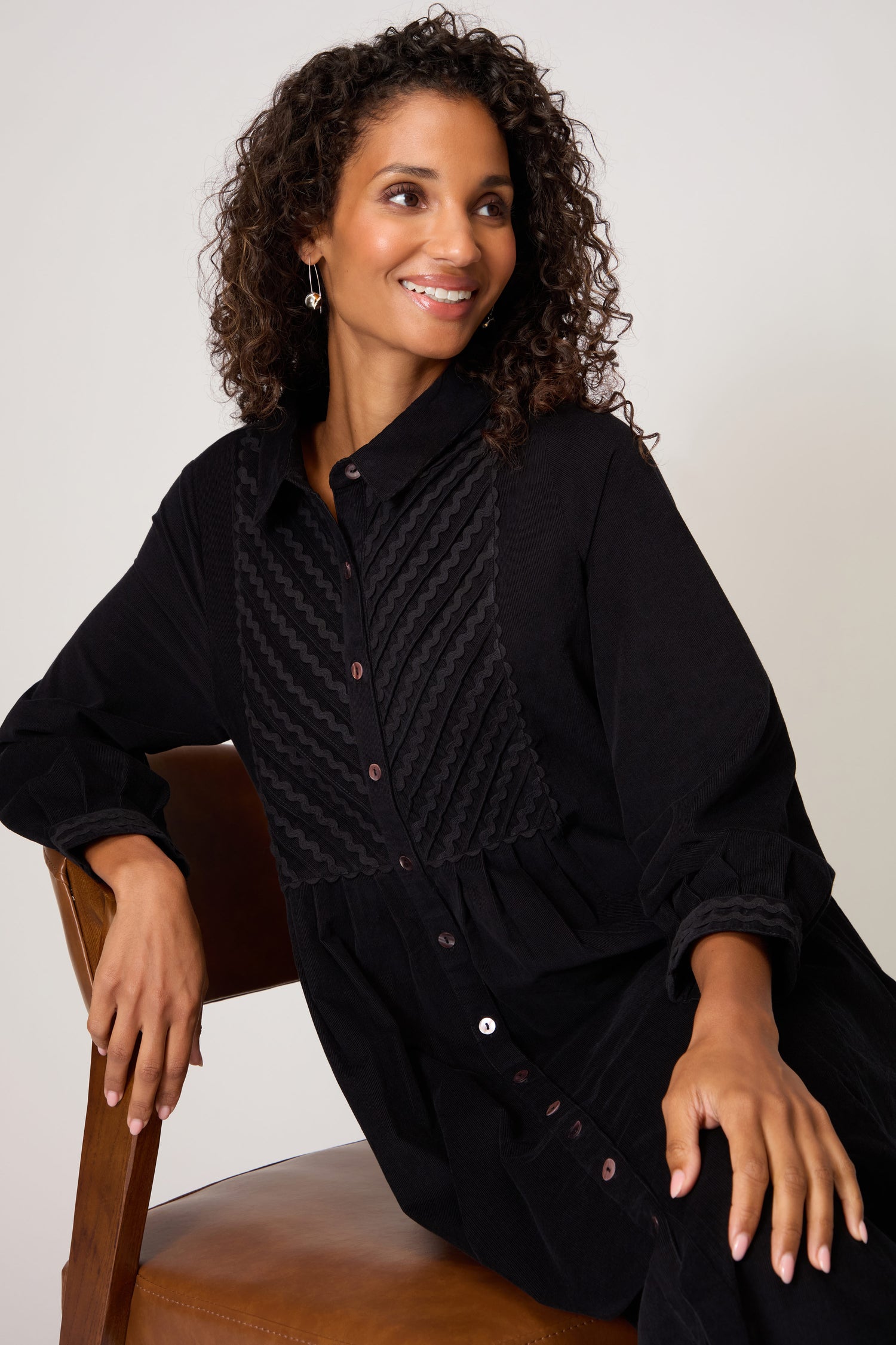 A woman with curly hair sits on a brown chair, wearing the Pinwale Cord Ric Rac Detail Dress, a classic-fit black A-line style with textured front detailing, as she looks off to the side and smiles.