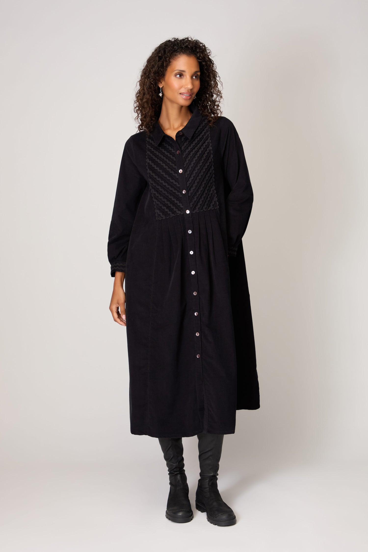 A woman stands against a plain background wearing the Pinwale Cord Ric Rac Detail Dress, a classic fit long black button-down cord dress with textured front panel, paired with black pants and boots.