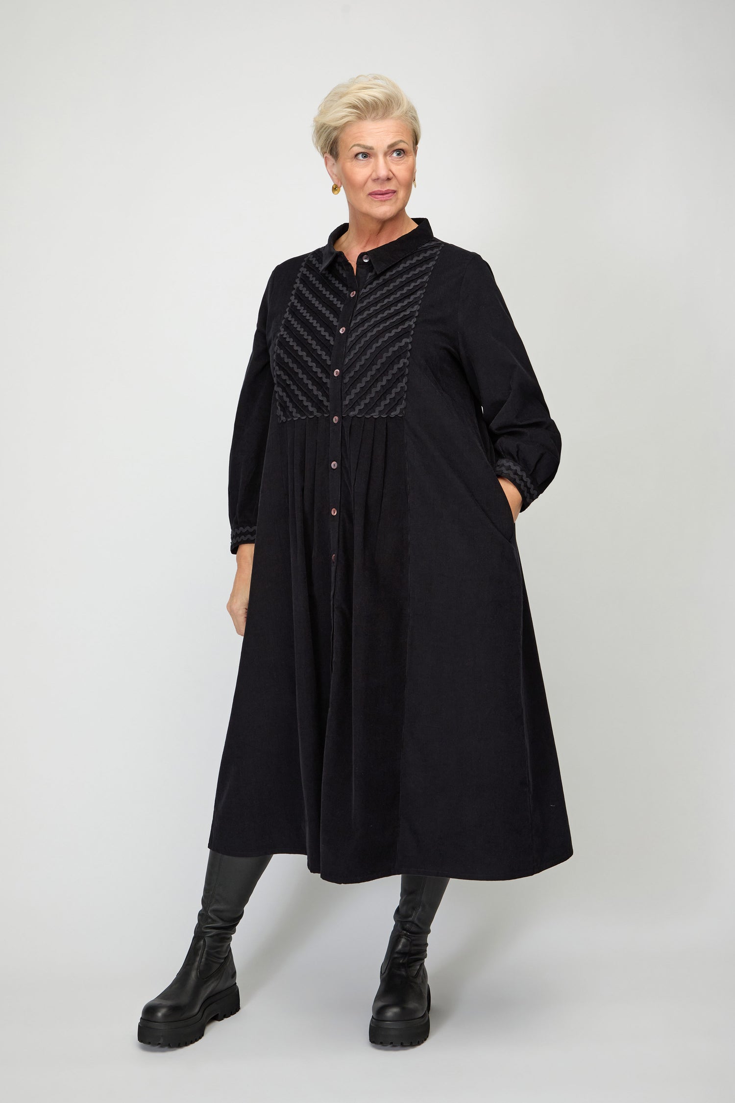 A woman with short blonde hair wears the Pinwale Cord Ric Rac Detail Dress, styled with black leggings and boots, and poses against a plain light background.