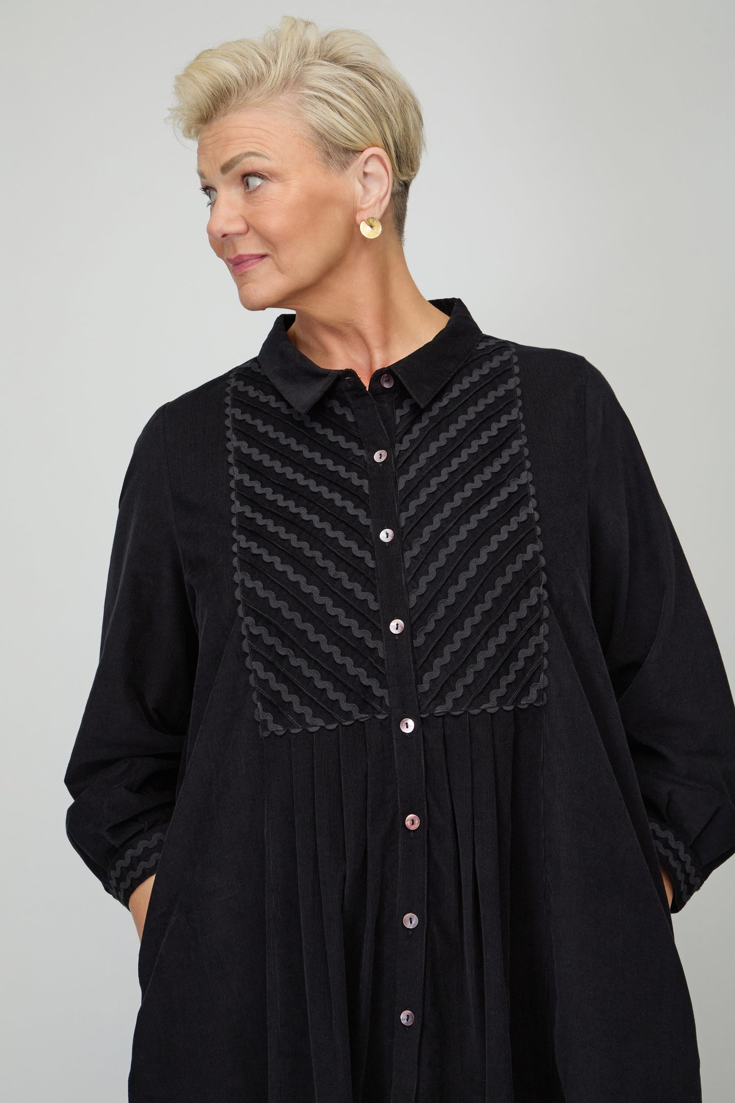 A person with short blond hair and gold earrings stands with hands in pockets, wearing the Pinwale Cord Ric Rac Detail Dress—a black A-line style featuring textured patterns—and looks to the side.