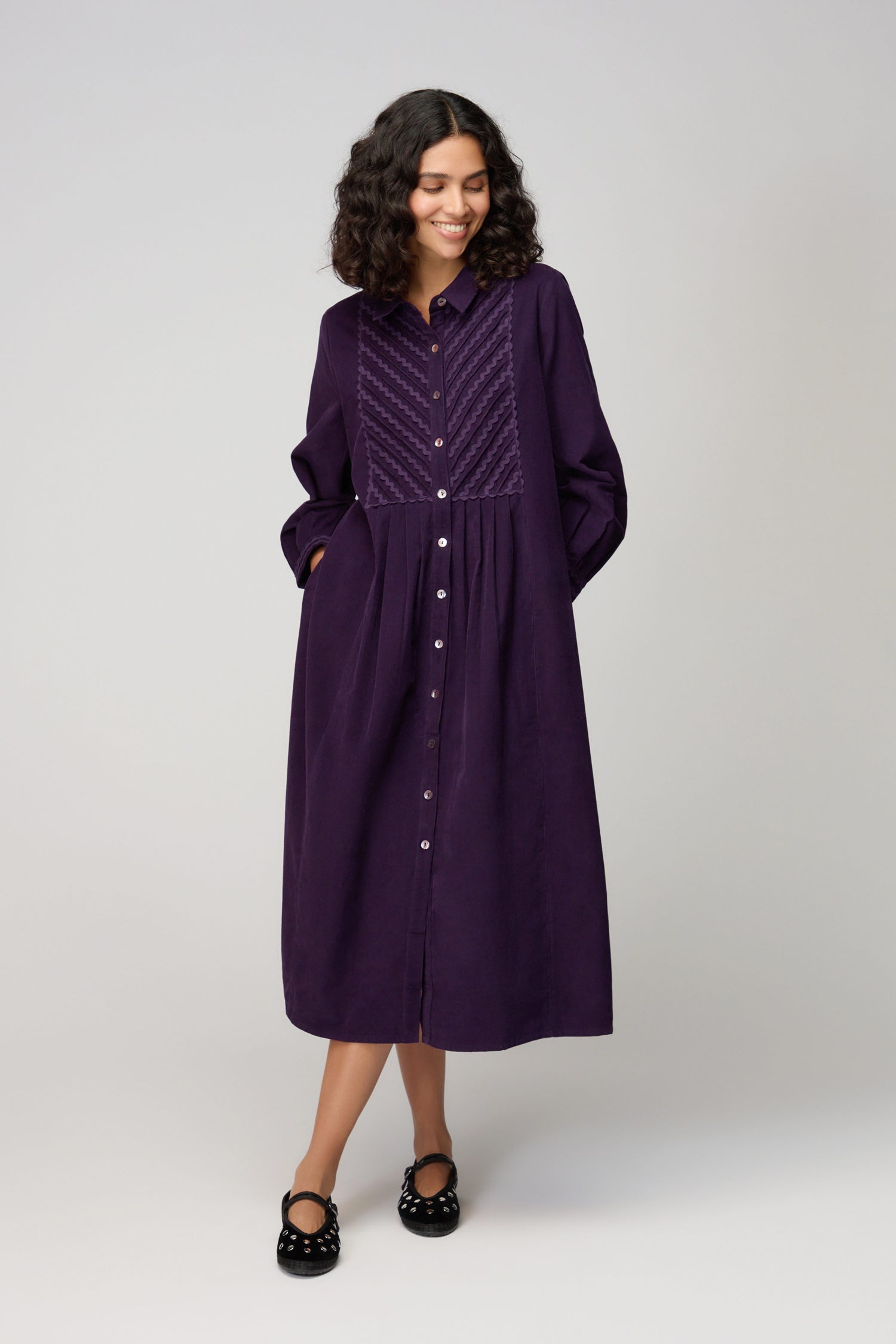A woman wears the Pinwale Cord Ric Rac Detail Dress—a long-sleeved, classic-fit, purple button-down dress with chest texture—paired with black shoes, standing against a light grey background.