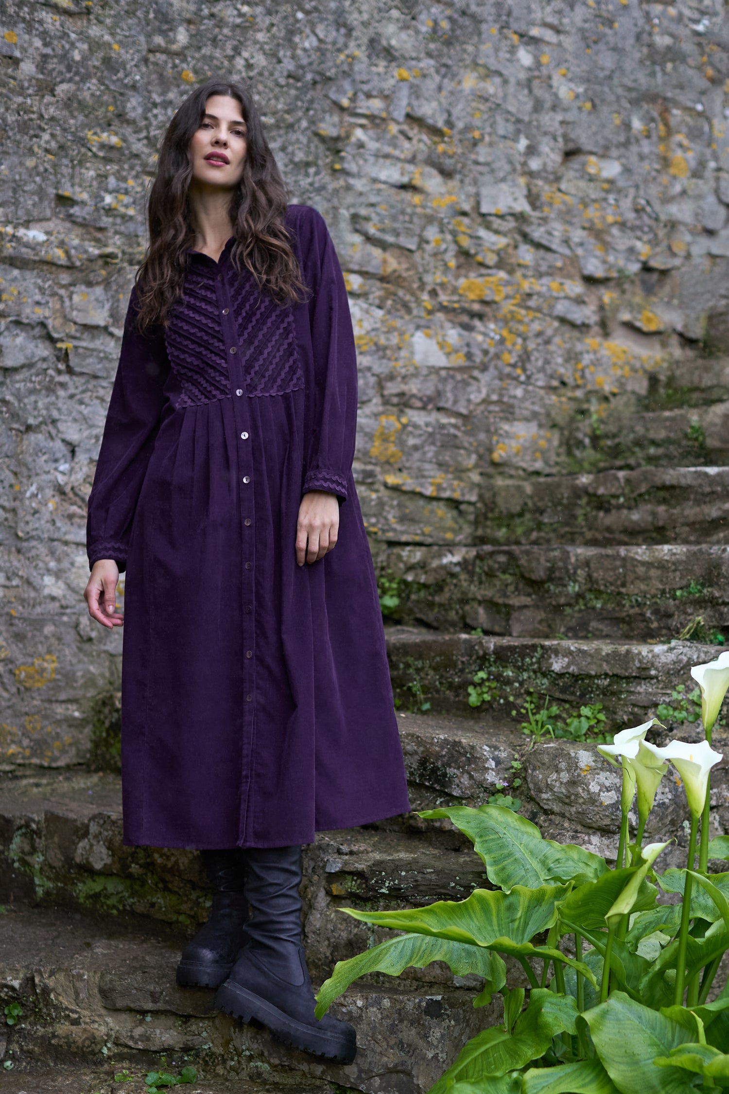 A woman in a Pinwale Cord Ric Rac Detail Dress and black boots stands on stone steps near white calla lilies, with a mossy stone wall behind her.