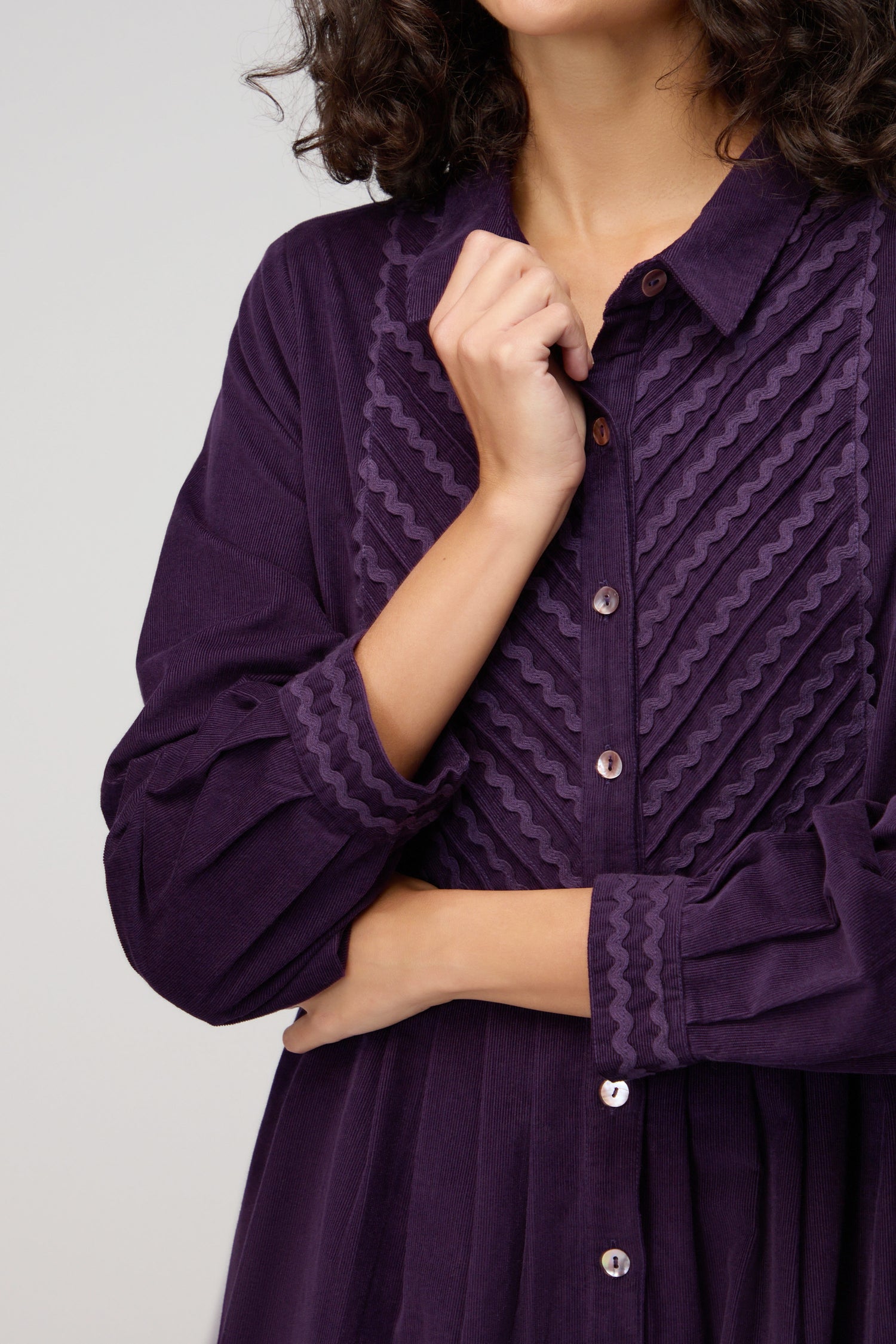 A woman wears the Pinwale Cord Ric Rac Detail Dress—a classic fit, long-sleeved, button-up purple dress with textured zigzag patterns—standing with one arm crossed and the other touching her collar.