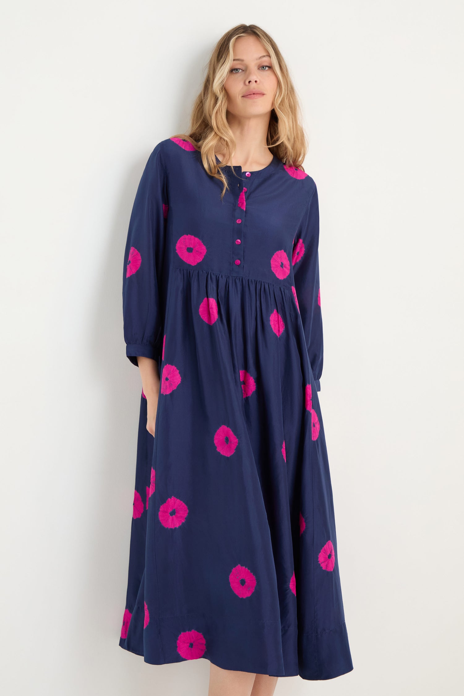 A woman wears the Shibori Spot Silk Dress, a long-sleeved, navy blue silk piece with pink circular patterns and a gathered waist, set against a plain light background.
