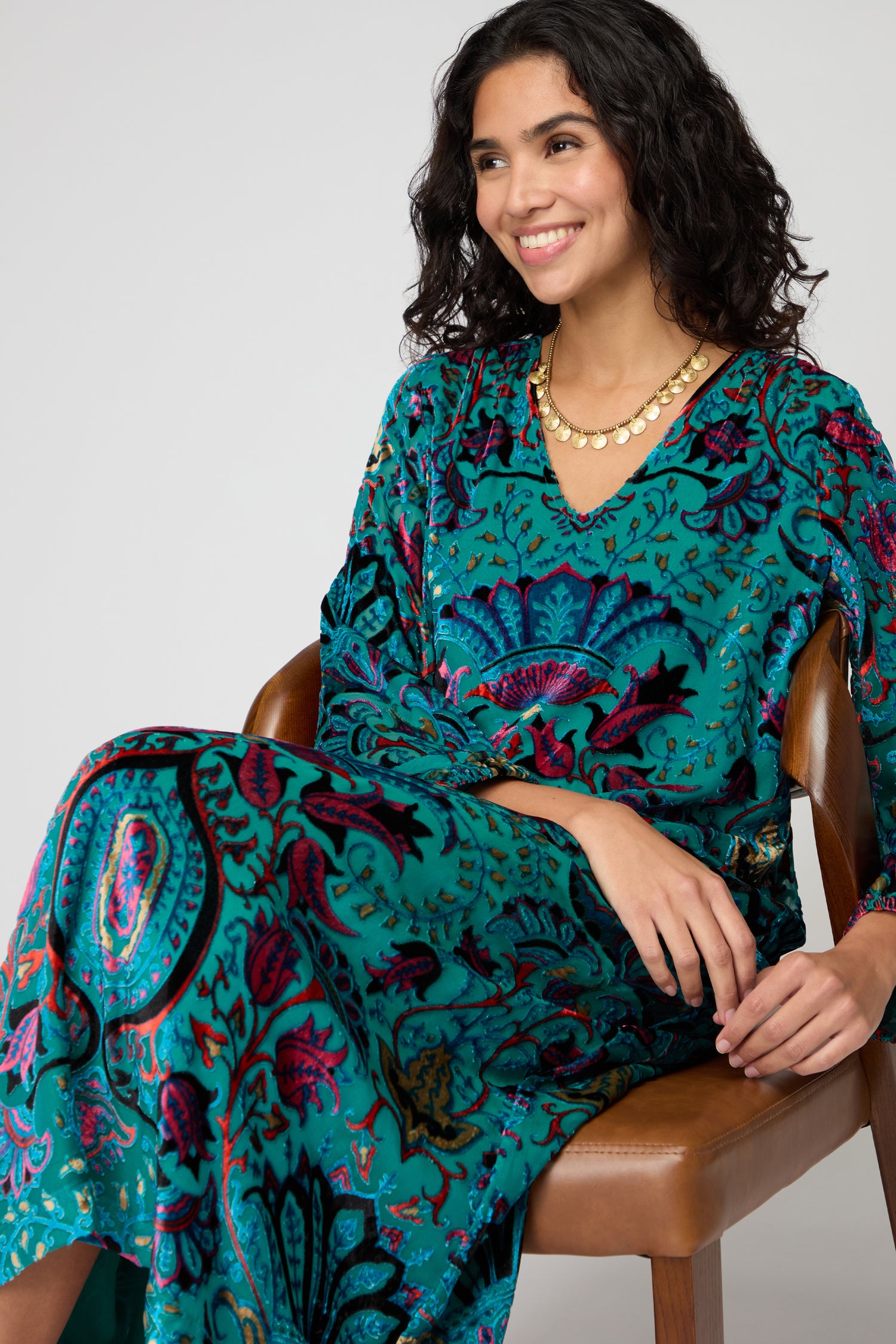 A woman with wavy dark hair sits on a brown chair, smiling and looking to the side, wearing the Winter Paisley Devore Dress—a velvet dress with a colorful floral pattern—paired with a gold necklace.