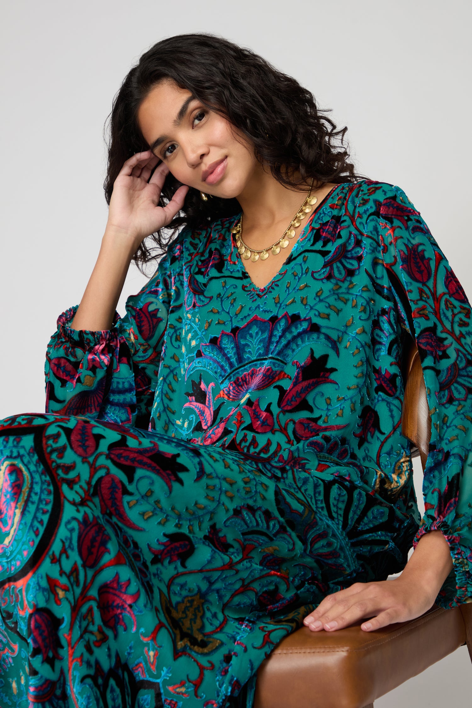 A woman with curly hair sits on a chair wearing the Winter Paisley Devore Dress in teal and magenta, paired with a gold necklace, resting her head on her hand against a plain background.