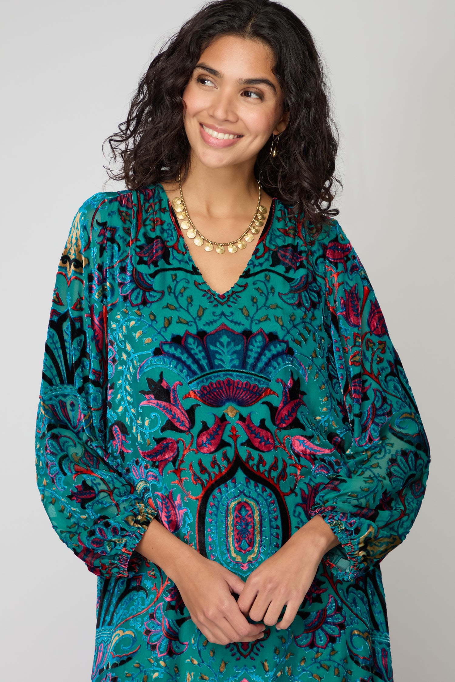 A woman wears the Winter Paisley Devore Dress featuring a teal and magenta paisley pattern and long sleeves, paired with a gold necklace and hoop earrings, smiling as she stands against a plain gray background.