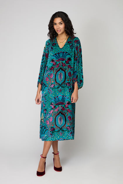 A woman stands against a light gray background wearing the Winter Paisley Devore Dress—a loose, teal paisley long-sleeve dress—paired with maroon heels and layered gold necklaces.
