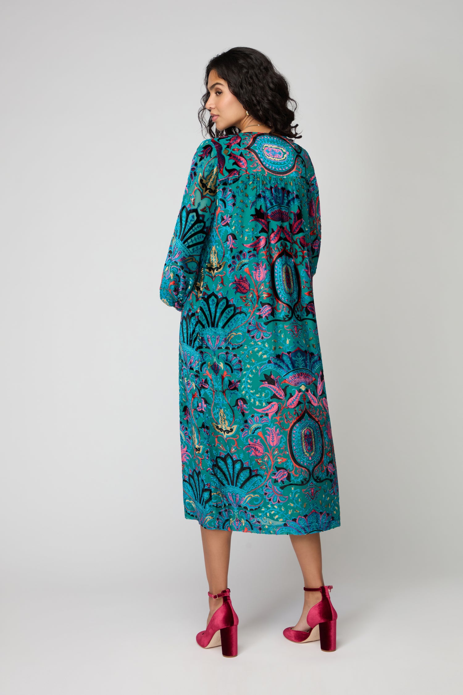A woman faces away, wearing the Winter Paisley Devore Dress in teal with a vibrant floral paisley pattern, paired with bright pink high heels. Ideal as a winter evening dress for stylish events.
