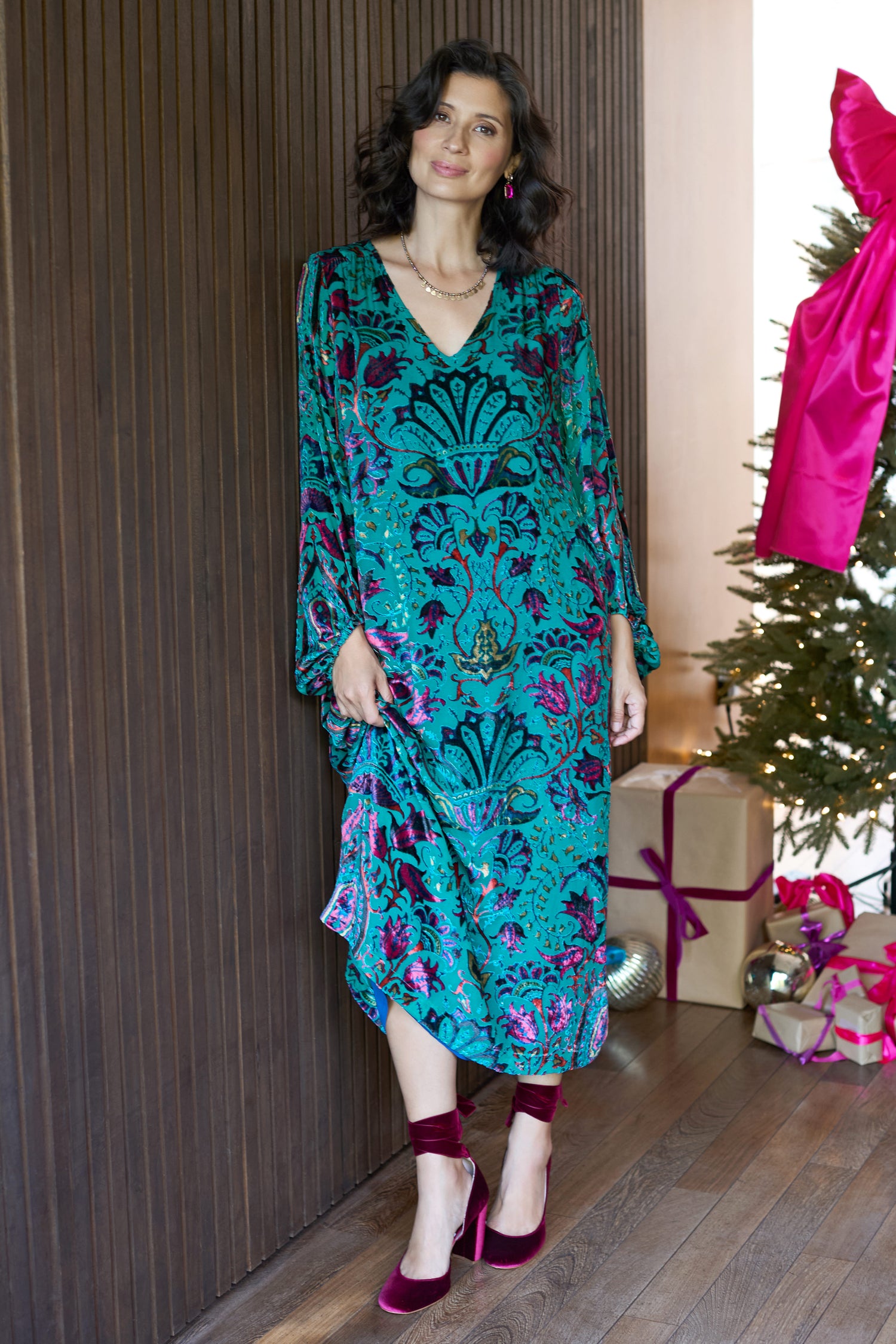 A woman wearing the Winter Paisley Devore Dress stands indoors beside a dark wooden wall and a decorated Christmas tree with wrapped gifts.