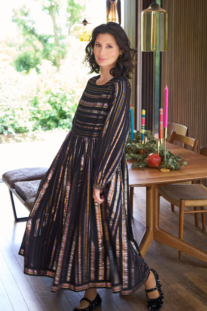 A woman in the Multi Stripe Lurex Dress stands indoors near a wooden dining table decorated with candles and greenery, sunlight streaming through large windows.