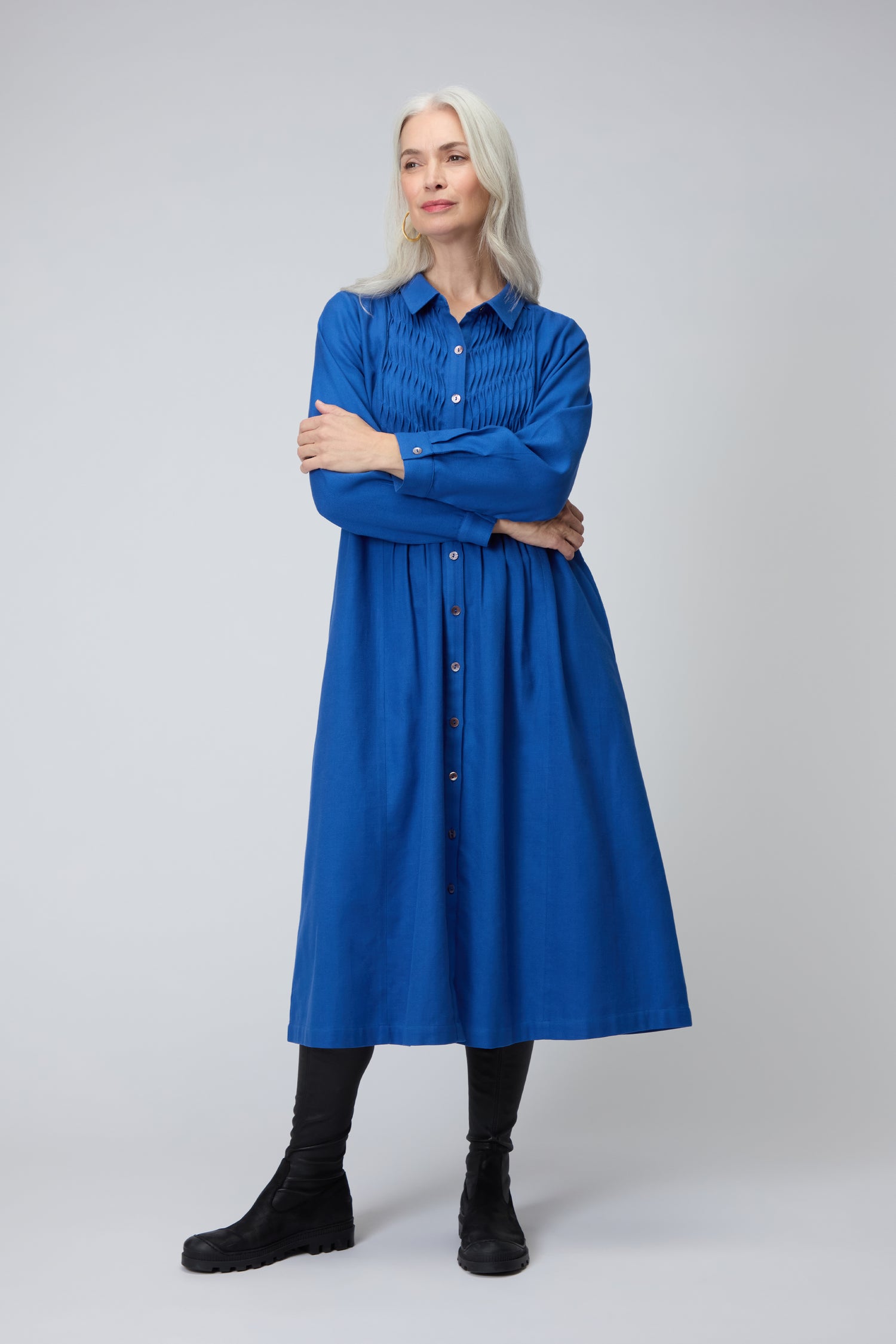 A woman with long gray hair stands with arms crossed against a plain background, wearing the Brushed Cotton Pintuck Detail A-line Dress paired with black leggings and shoes.