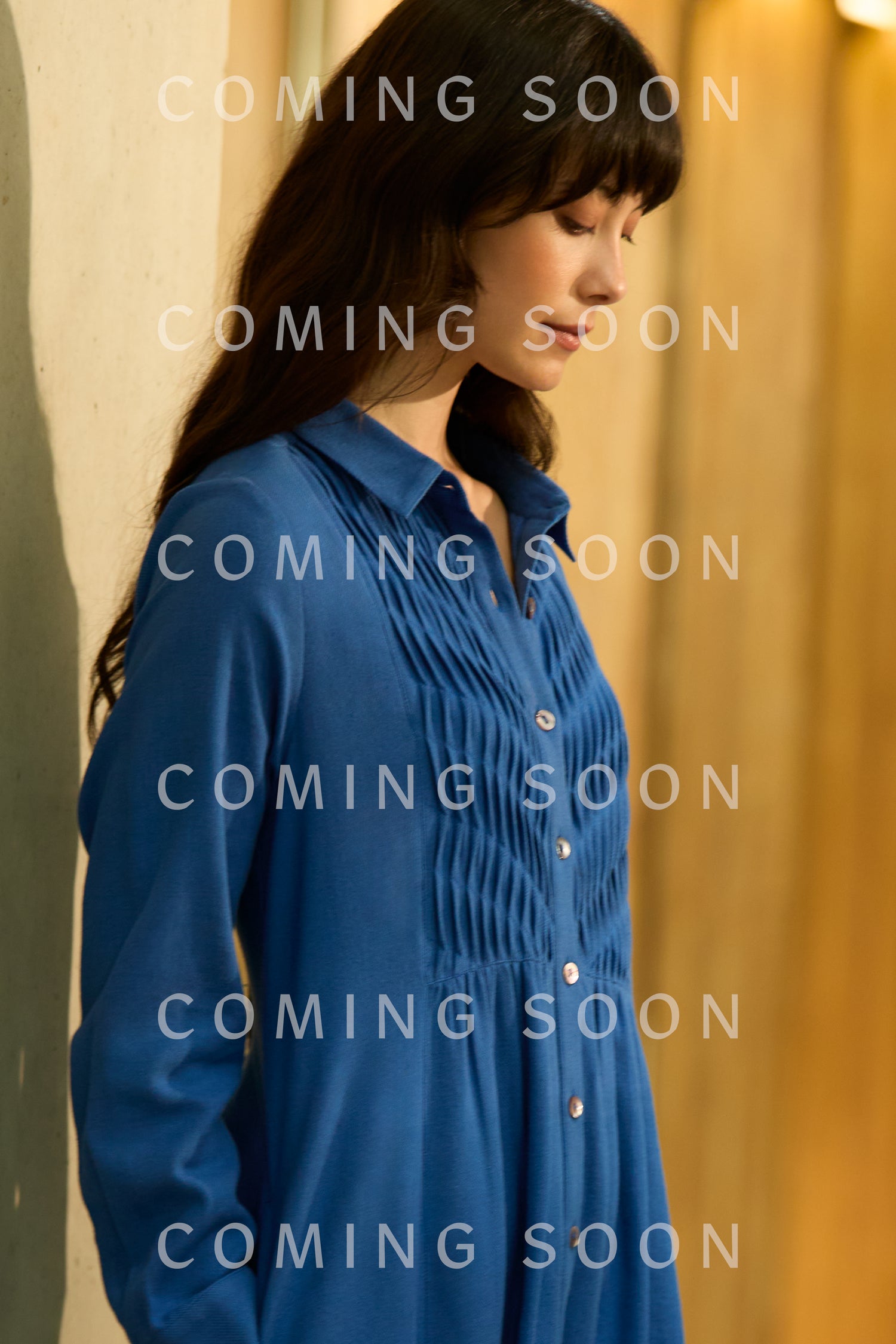 A woman in a blue Brushed Cotton Pintuck Detail A-line Dress stands indoors, looking down. "COMING SOON" is repeatedly overlaid on the image.