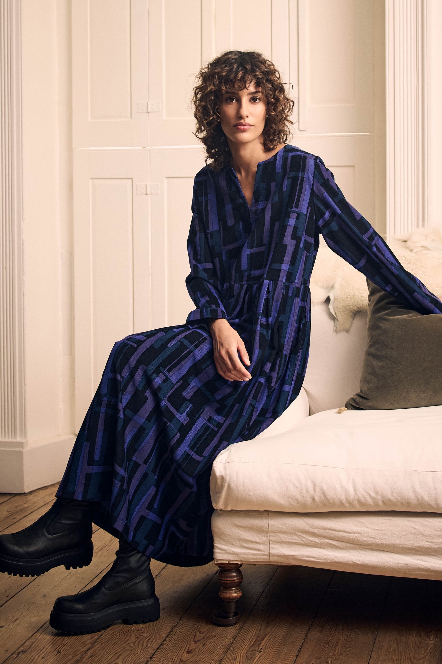 A woman wearing a Layered Geometric Fine Cord Flared Dress with a blue and purple grid pattern sits on a white sofa, paired with black ankle boots.