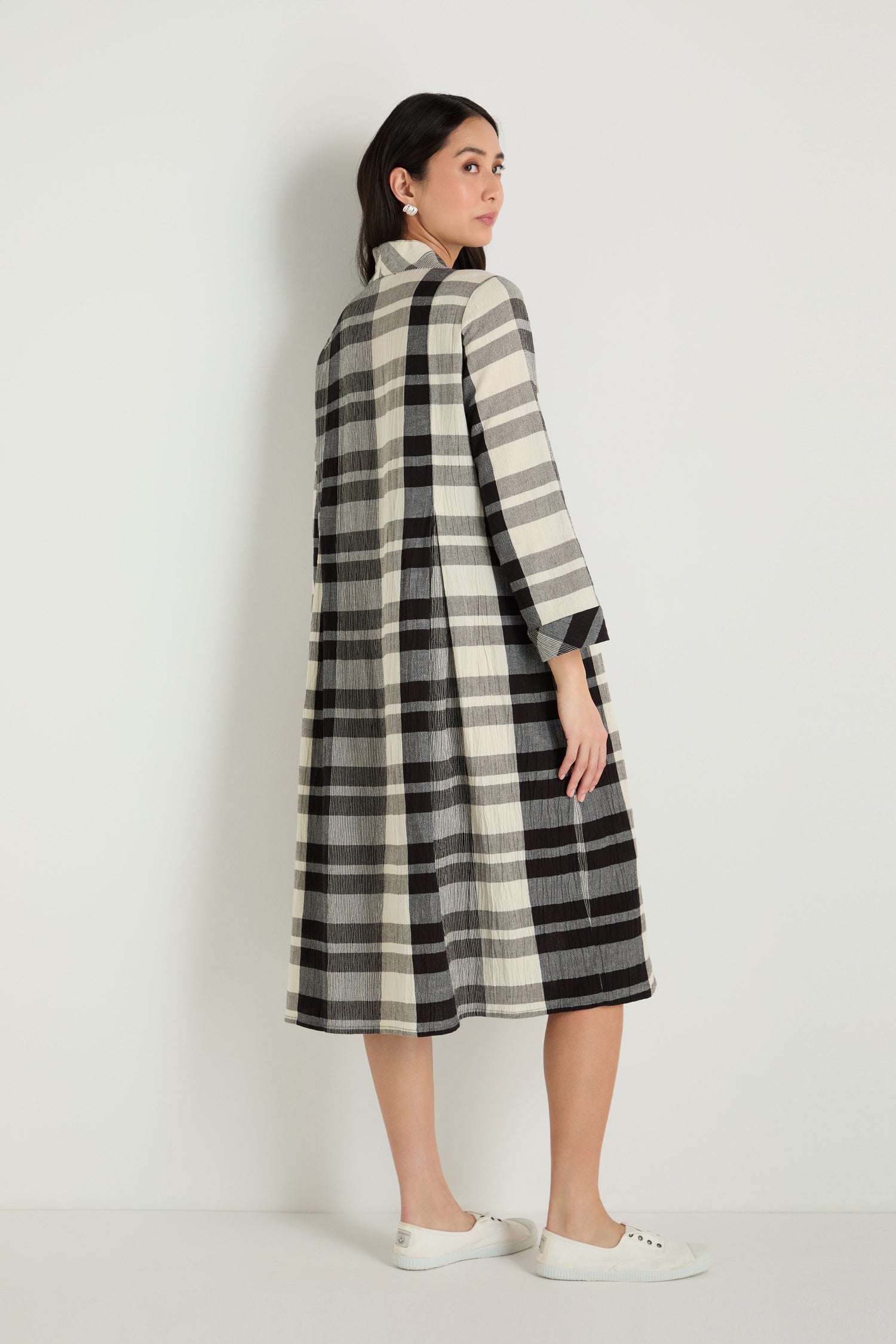 A woman stands facing slightly away, wearing a Monochrome Crinkle Check Dress with long sleeves and black slip-on shoes.