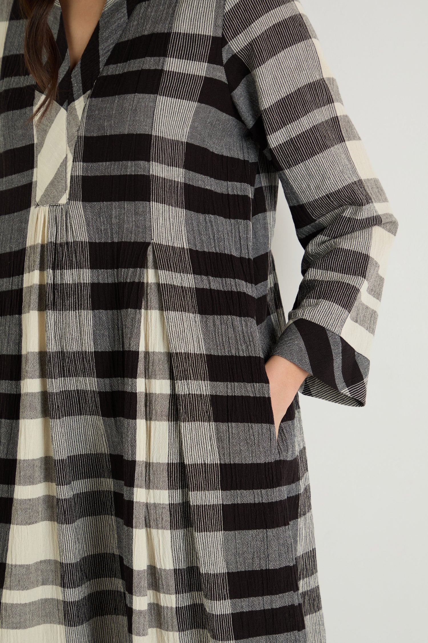 A person wearing a long-sleeved Monochrome Crinkle Check Dress in black, gray, and cream plaid with one hand in a side pocket.