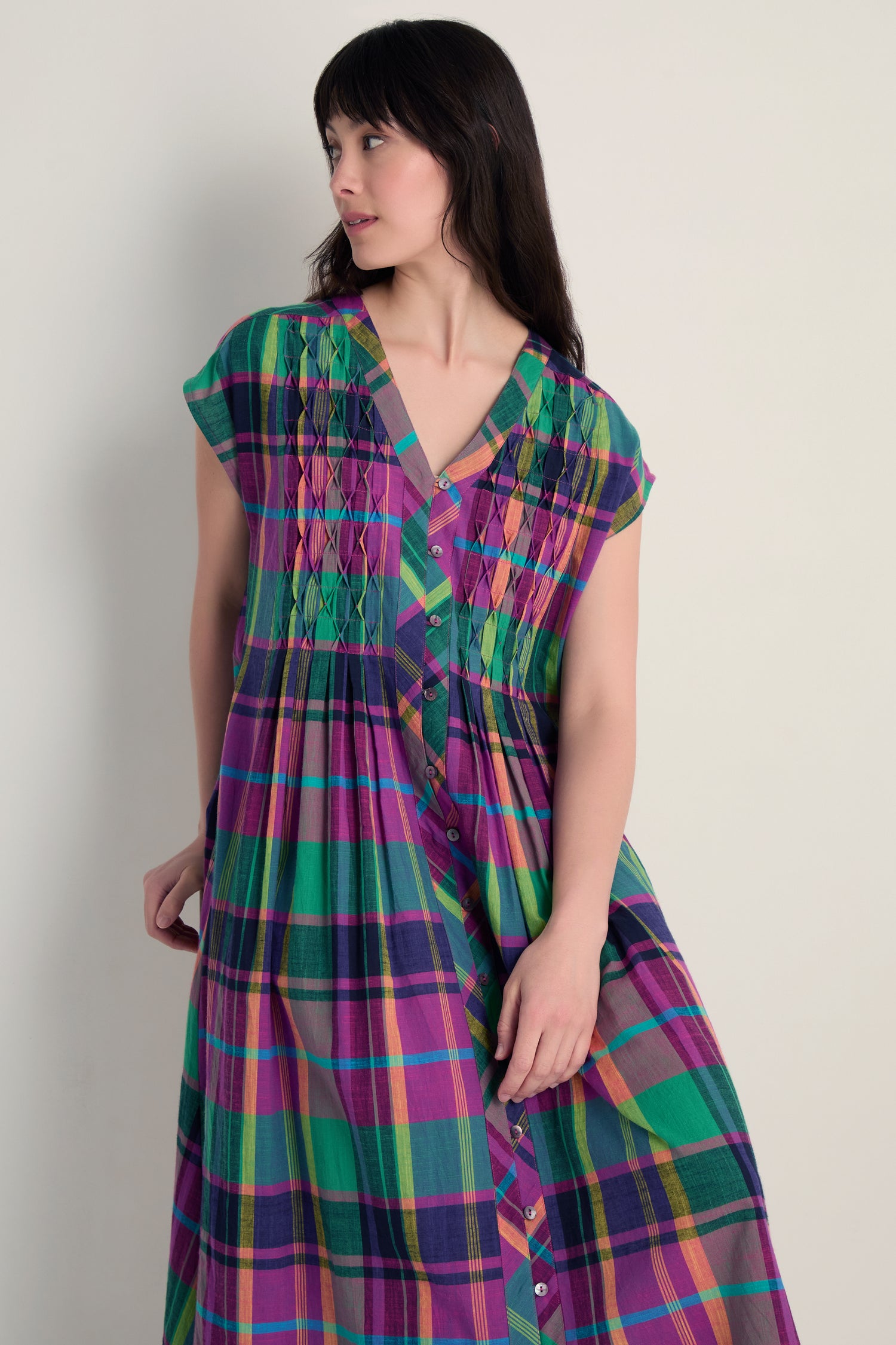 A woman models the Madras Check Dress, featuring colorful checks, a button-down front, and honeycomb pleats, set against a plain light-colored background.