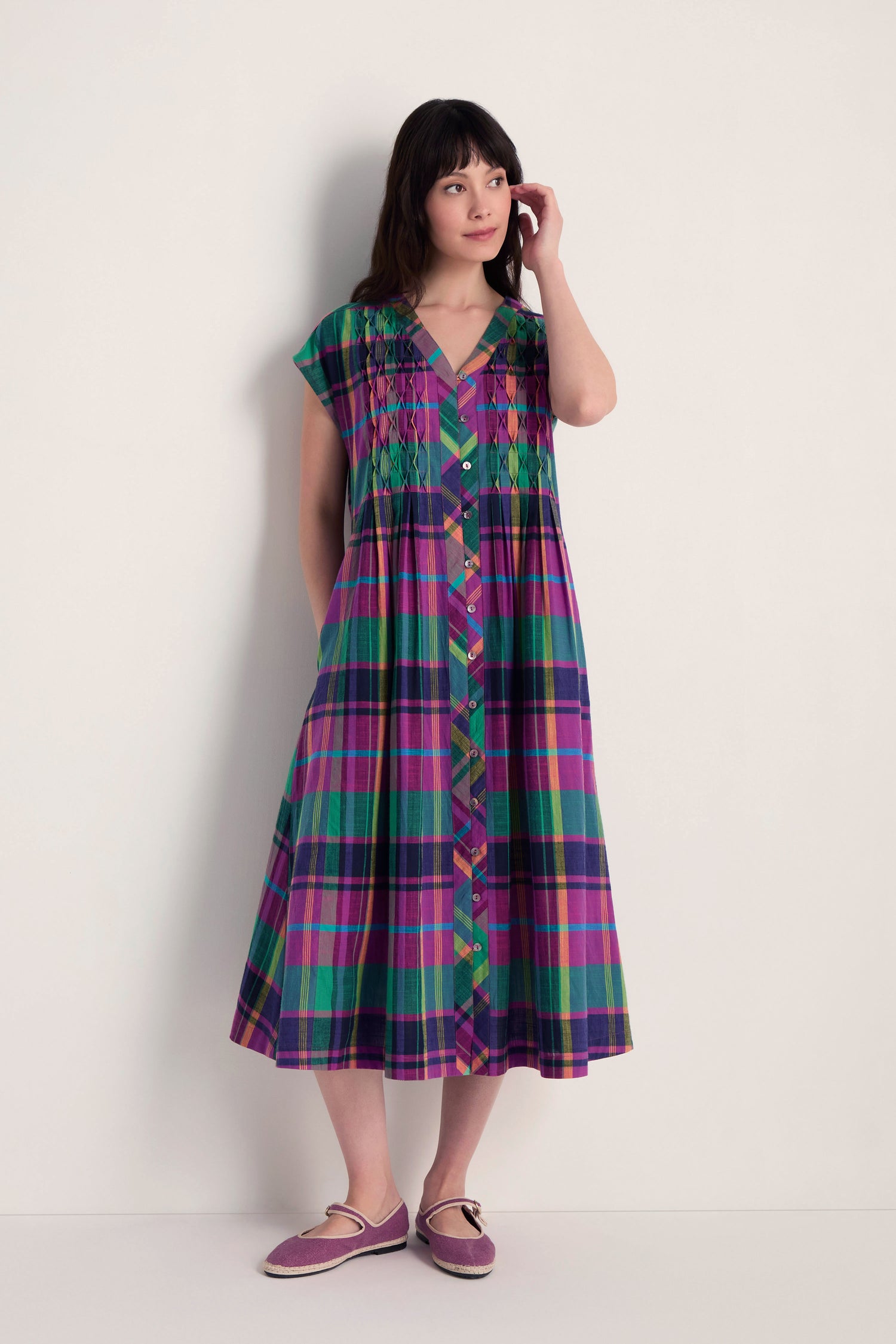 A woman stands against a plain wall wearing the Madras Check Dress featuring short sleeves and honeycomb pleated detailing, paired with purple slip-on shoes.