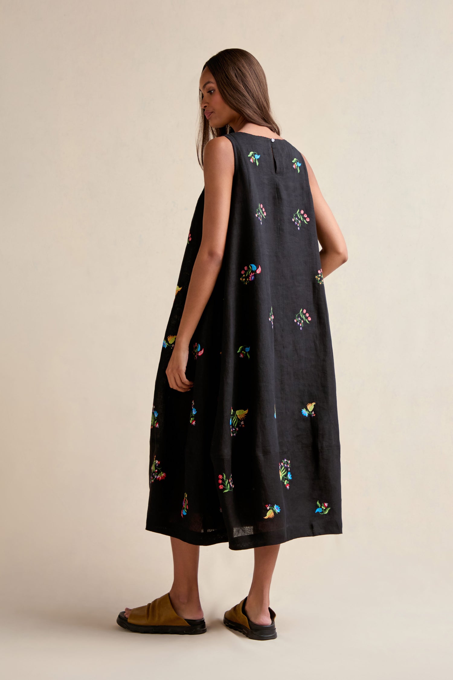 A woman faces away, wearing the Vintage Floral Embroidered Bubble Dress with colorful patterns and brown sandals, set against a plain light background.