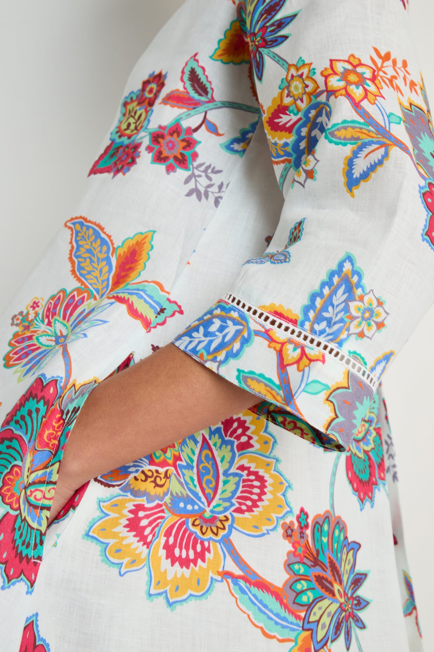 Close-up of a person wearing the Botanical Linen Flare Dress, a white linen piece adorned with colorful, intricate sarasa-inspired floral patterns. The person's hand rests partially inside one of the dress's side pockets.