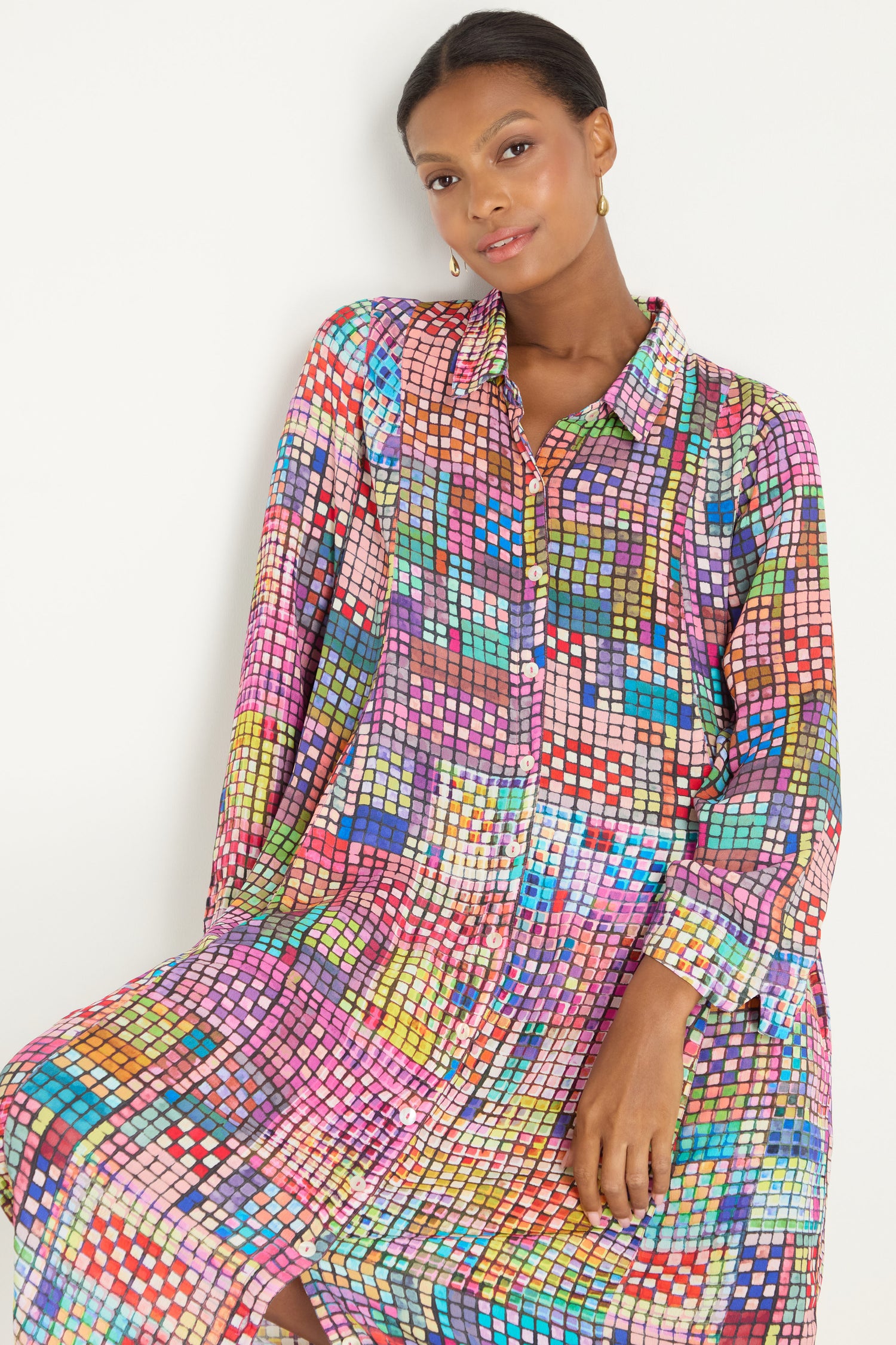 A woman wears the Coloured Glass Tiles Shirt Dress, a loose, long-sleeved dress featuring a colorful geometric pattern, standing against a plain light background.