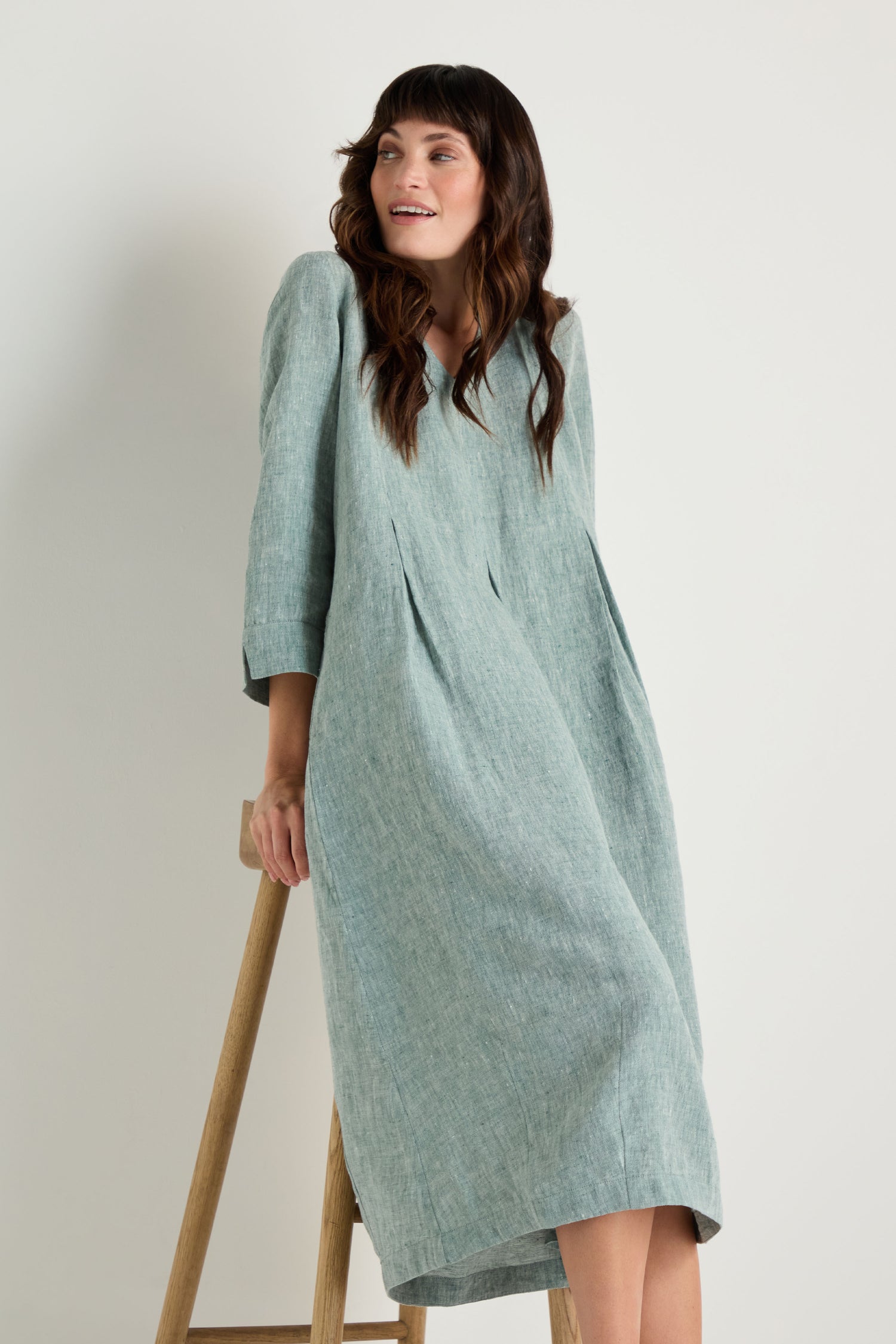 A woman in the Crinkle Linen Cross Dye Bubble Dress, featuring a light blue crinkle linen fabric and bubble silhouette, stands leaning on a wooden stool against a plain white background.