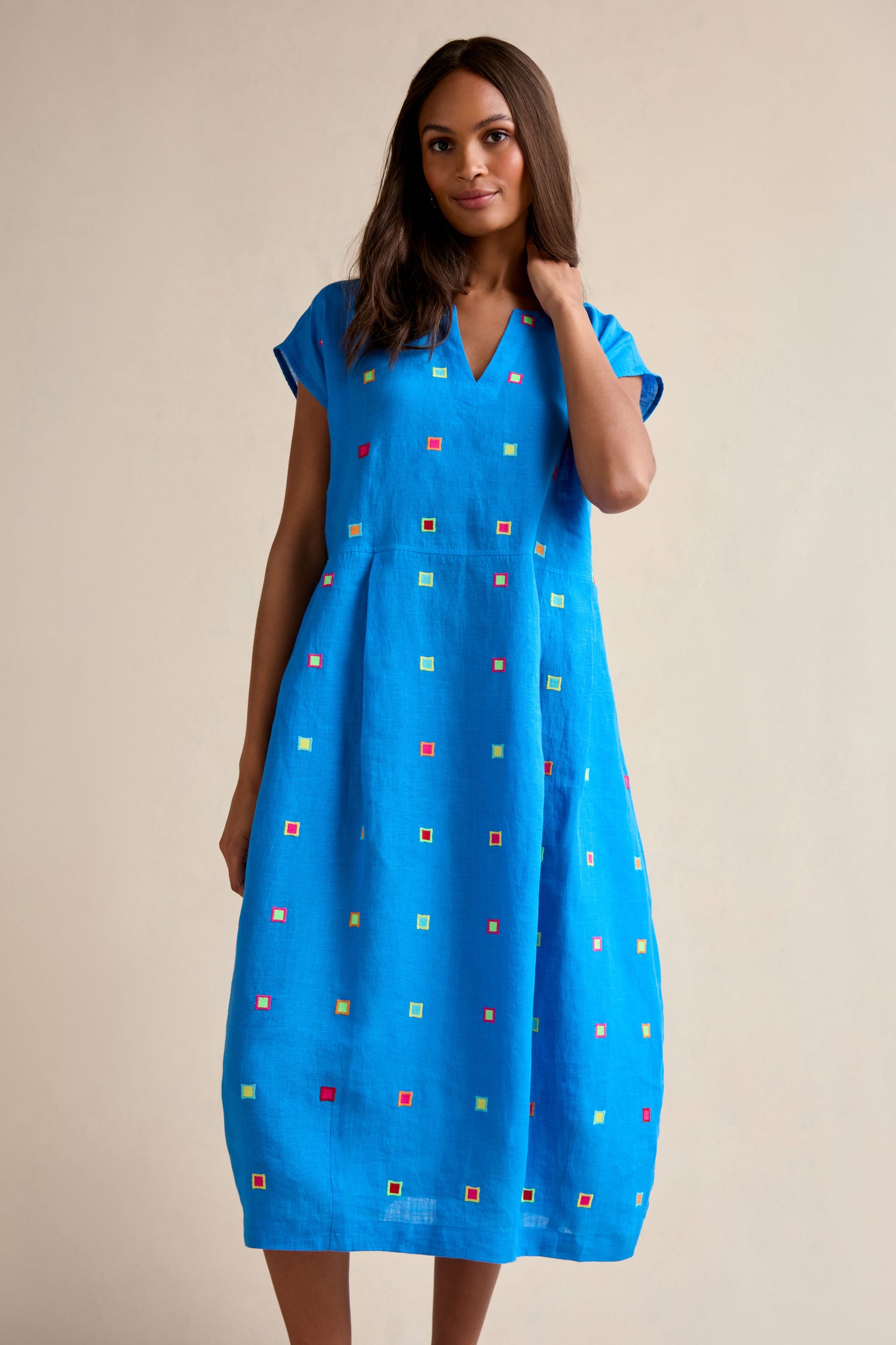 A woman stands against a neutral background wearing the Embroidered Squares Linen Bubble Dress, a blue, short-sleeve, midi-length linen dress adorned with colorful embroidered squares.