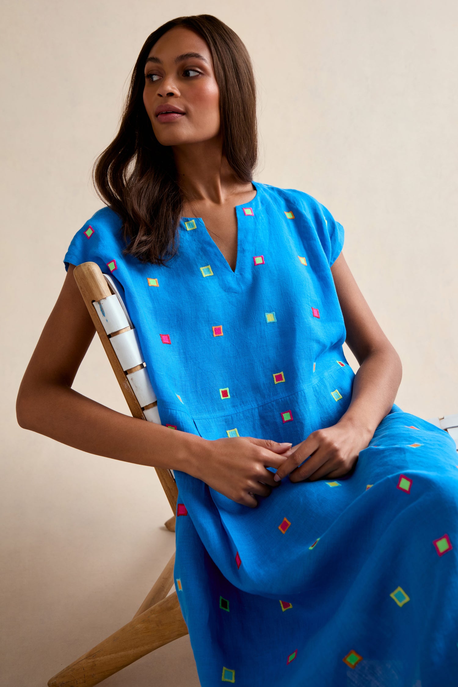 A woman sits in a wooden chair wearing the Embroidered Squares Linen Bubble Dress—a bright blue, short-sleeved, midi-length linen dress adorned with small, colorful square patterns.