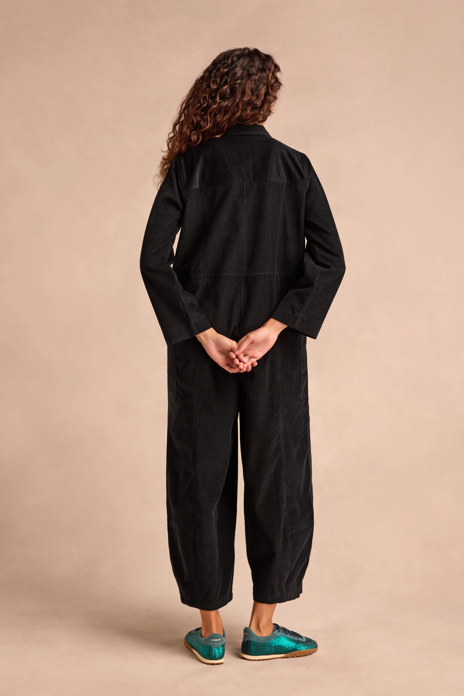 A person with long curly hair stands facing away, wearing the loose-fitting Pinwale Cord Jumpsuit—crafted from pure cotton with a bubble leg silhouette—paired with teal shoes and hands clasped behind their back against a plain beige background.