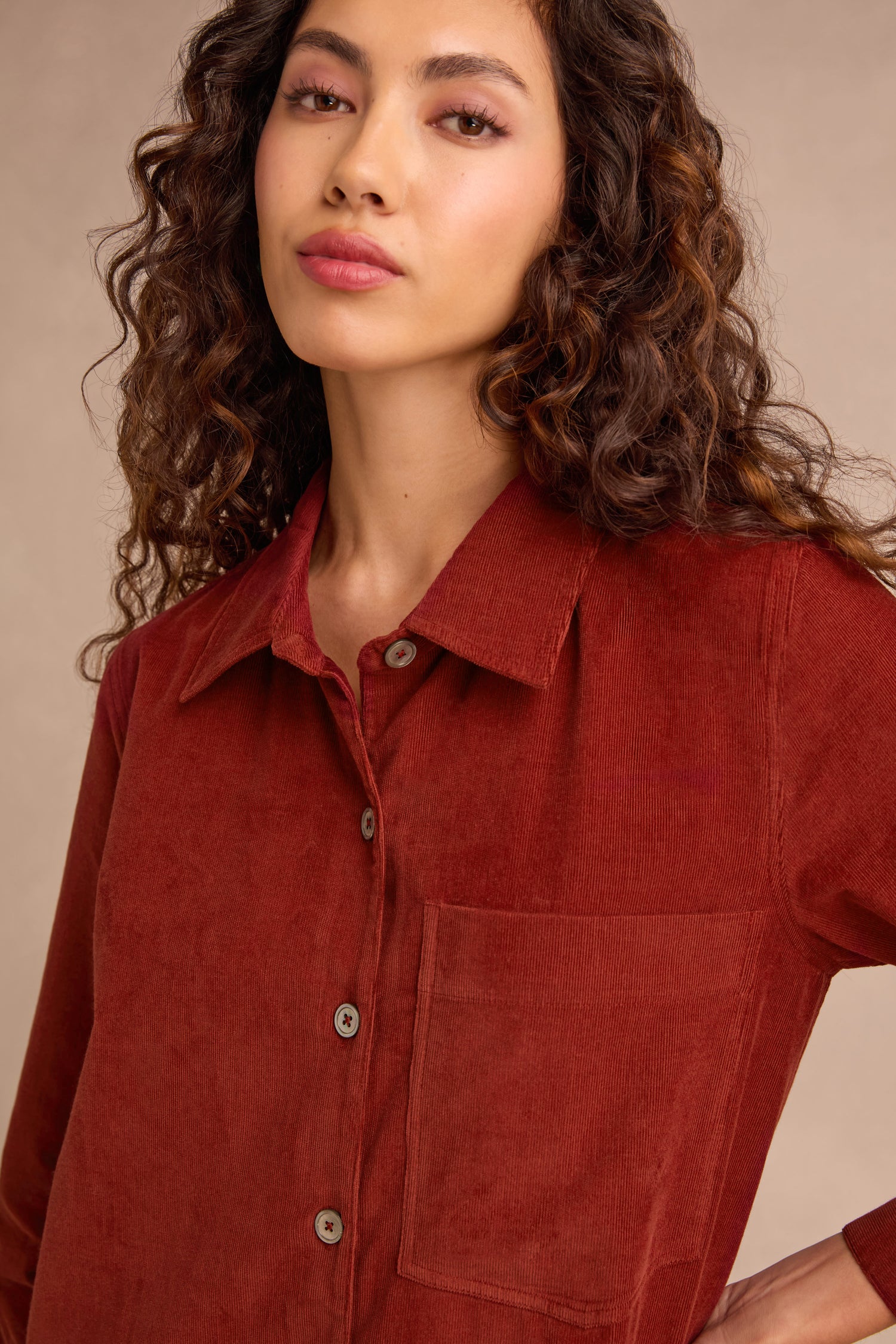 A woman with long curly hair wears the Conker Pinwale Cord Shirt Dress, featuring a rust hue and a large front pocket, standing against a beige background.