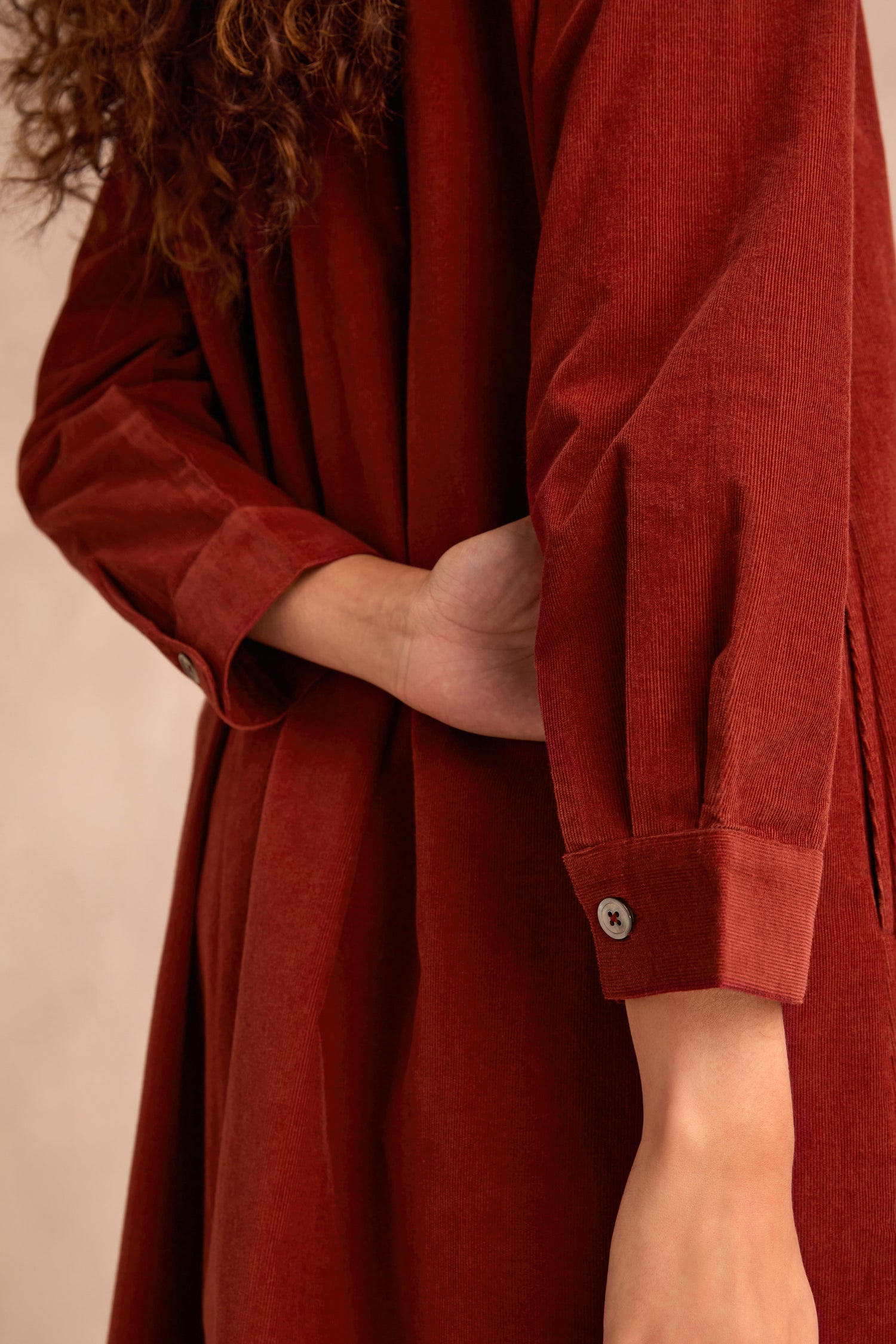 A person wearing the Conker Pinwale Cord Shirt Dress with one arm bent behind their back, highlighting a close-up of the red cord fabric and buttoned cuff.