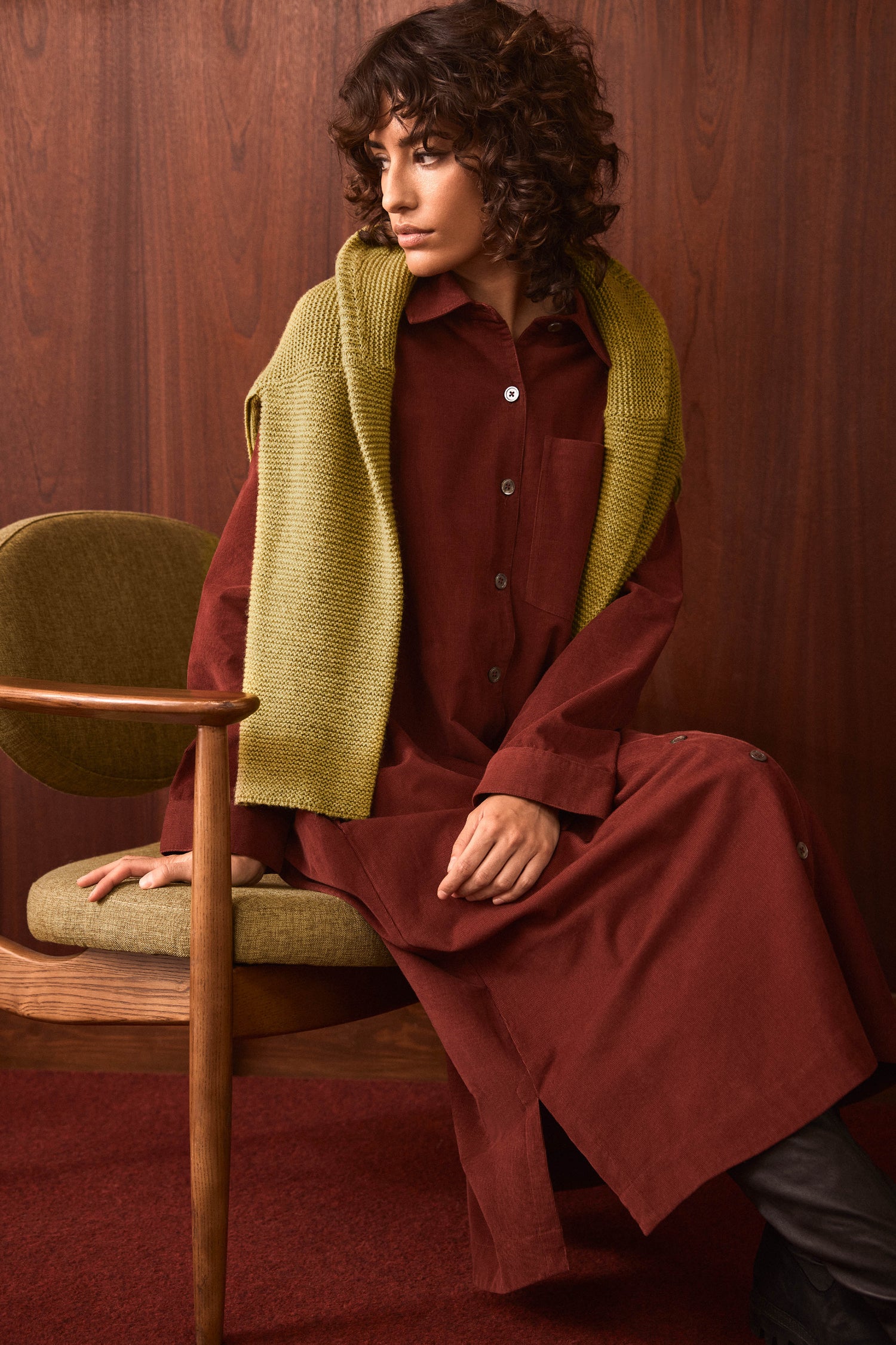 A woman wearing the Conker Pinwale Cord Shirt Dress in rich maroon sits on a green chair, with a green knit sweater draped over her shoulders, against a wood-paneled wall.