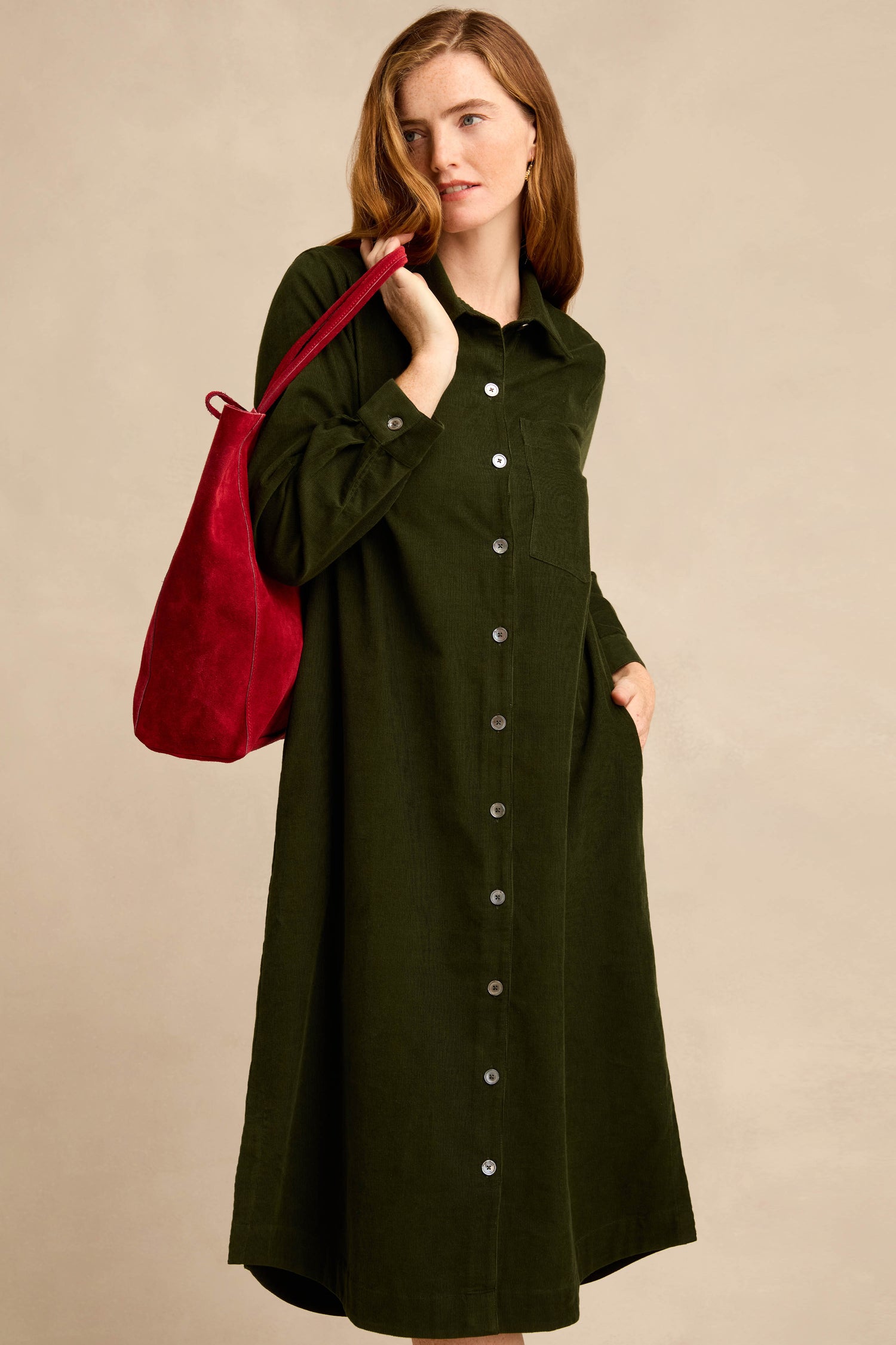 A woman stands against a plain beige background, wearing the Forest Pinwale Cord Shirt Dress—a long-sleeve, button-down, dark green corduroy dress with a breast pocket—while carrying a red shoulder bag.