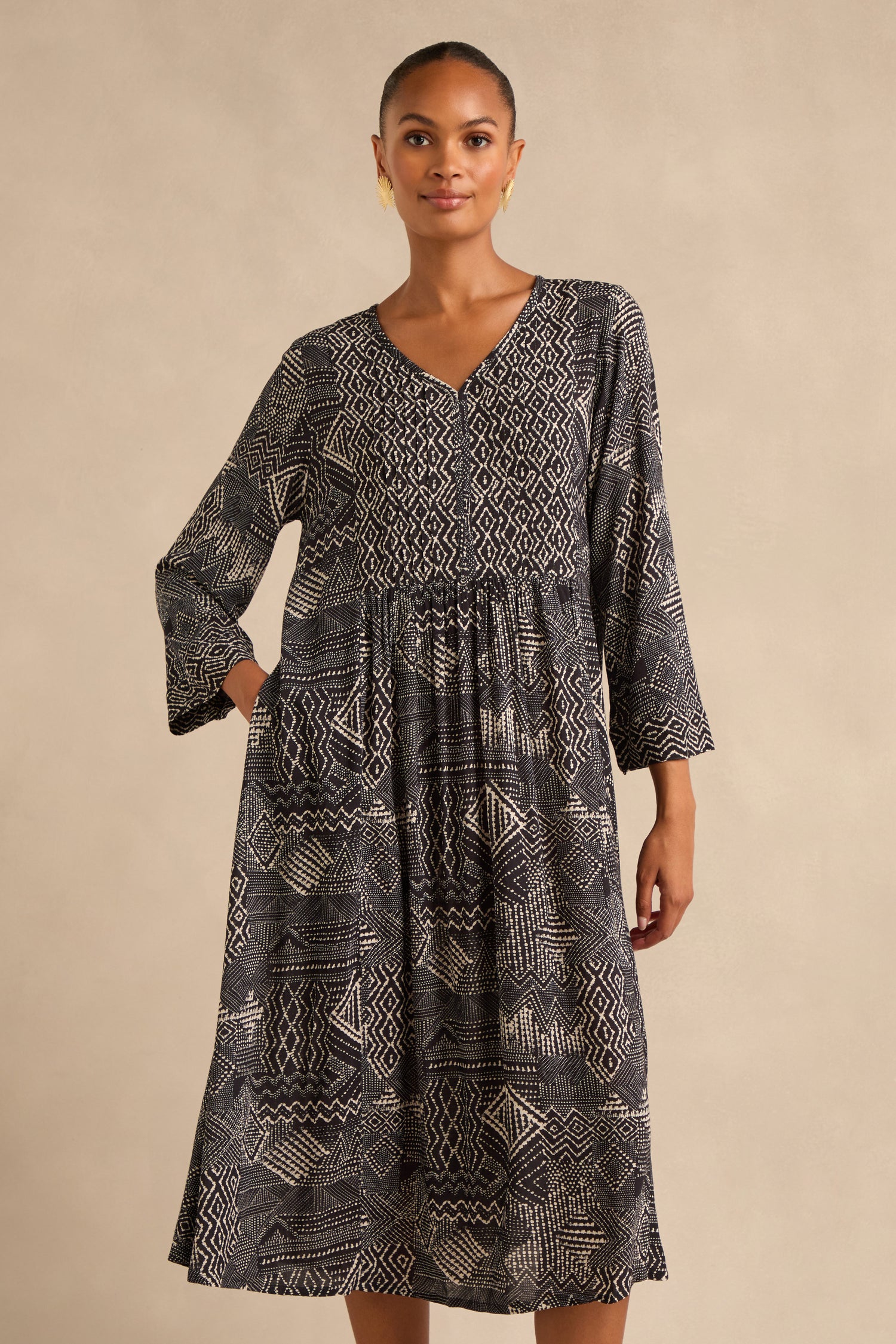 A woman stands against a neutral background, wearing the Mono Patchwork Ikat Dress—an A-line silhouette with long sleeves, a V-neckline, a gathered waist, and a striking black and white pattern.
