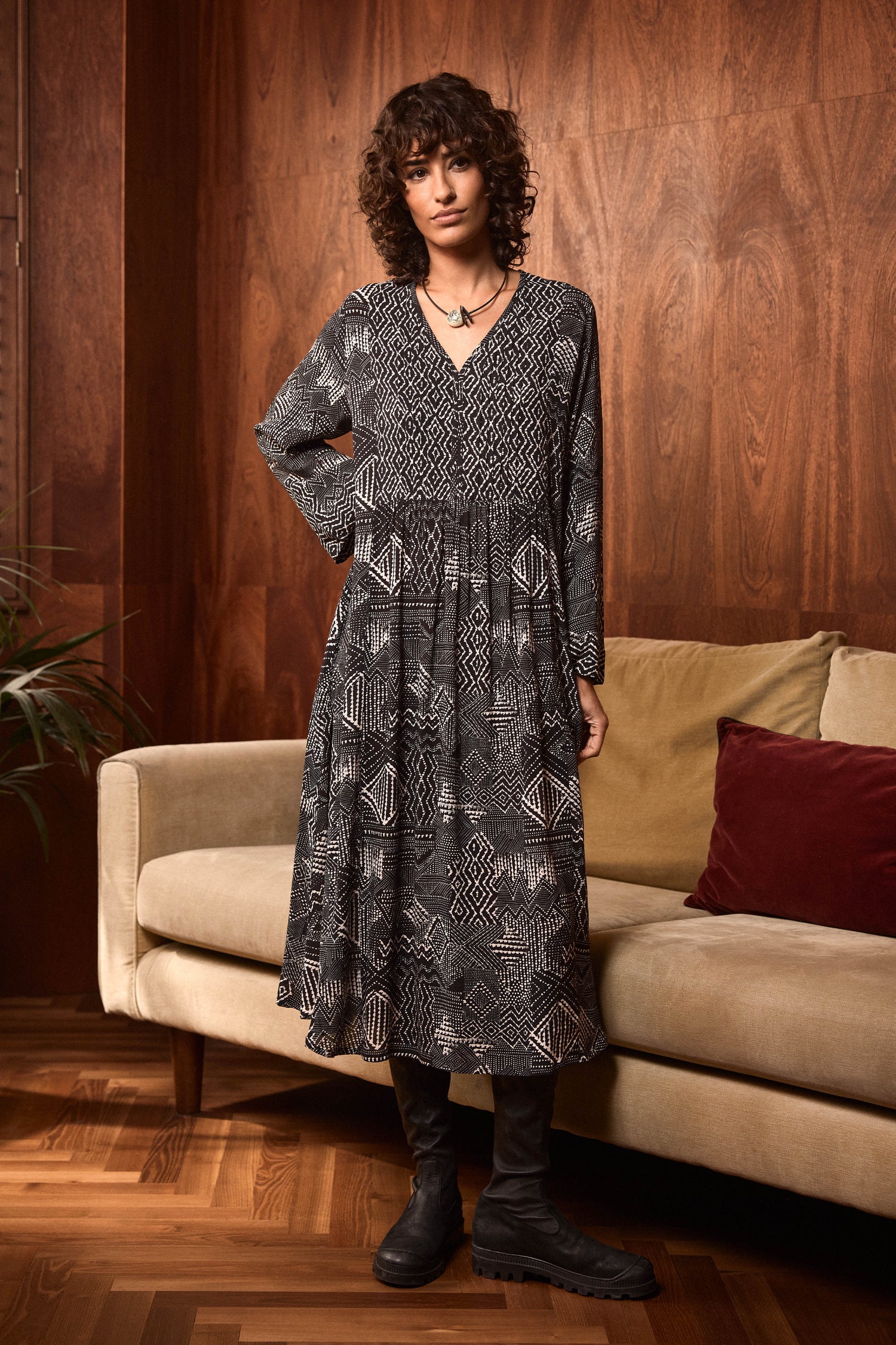 A person stands indoors wearing the Monochrome Batik Print Dress, which features long sleeves and an elegant A-line silhouette, paired with black boots. They are positioned in front of a beige sofa adorned with red and tan pillows, set against a wooden wall background.