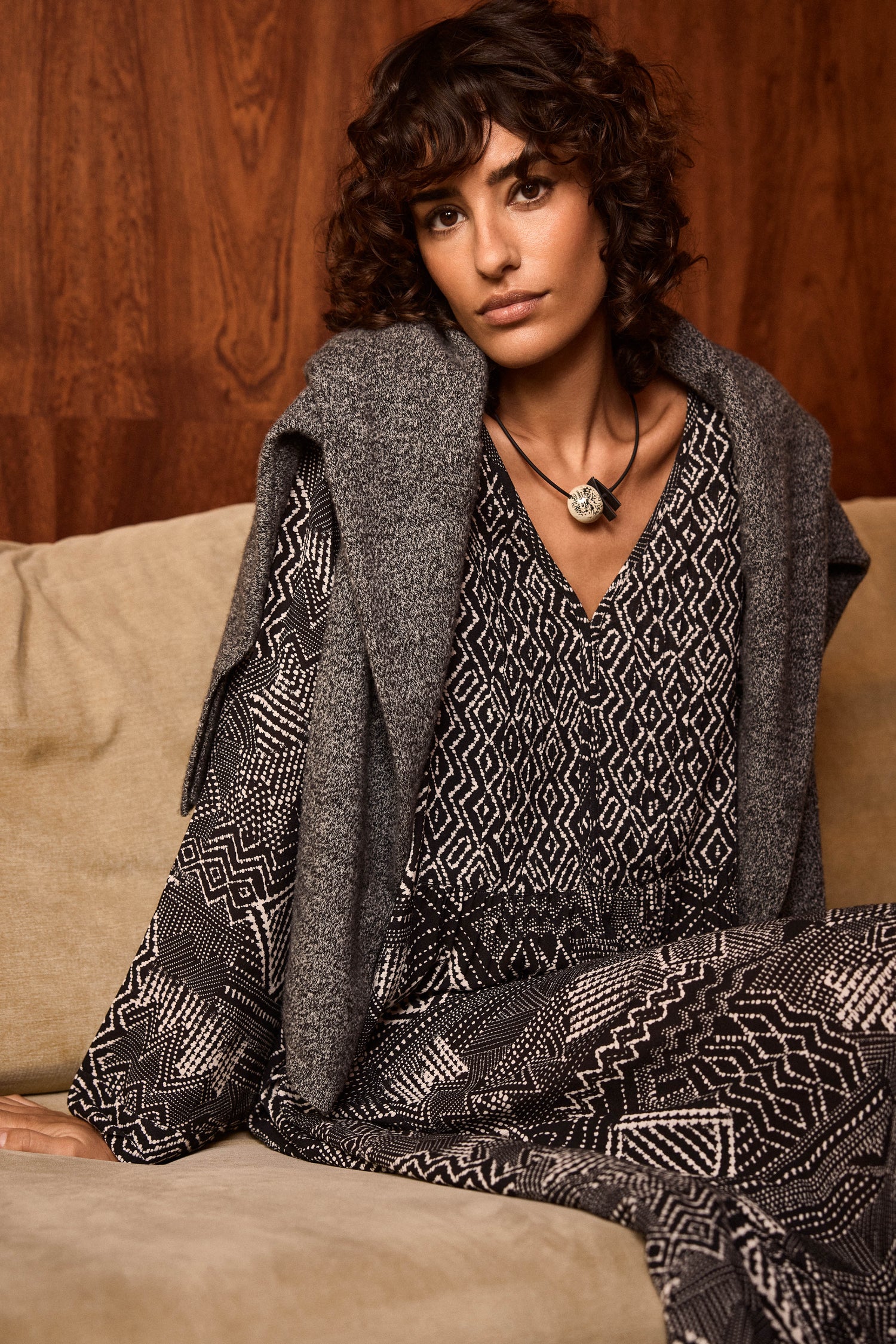 A woman with curly hair sits on a beige sofa, wearing the Monochrome Batik Print Dress in an A-line silhouette, a gray sweater draped over her shoulders, and a pendant necklace, with a wooden wall as the background.