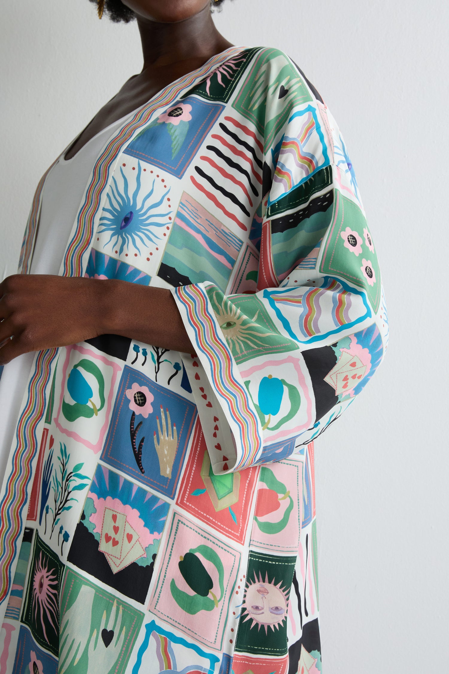 Wearing the Mystical Collage Kimono with its colorful, abstract, and nature-inspired patterns, someone poses by a plain light wall—ideal for those who love a free-spirited look.