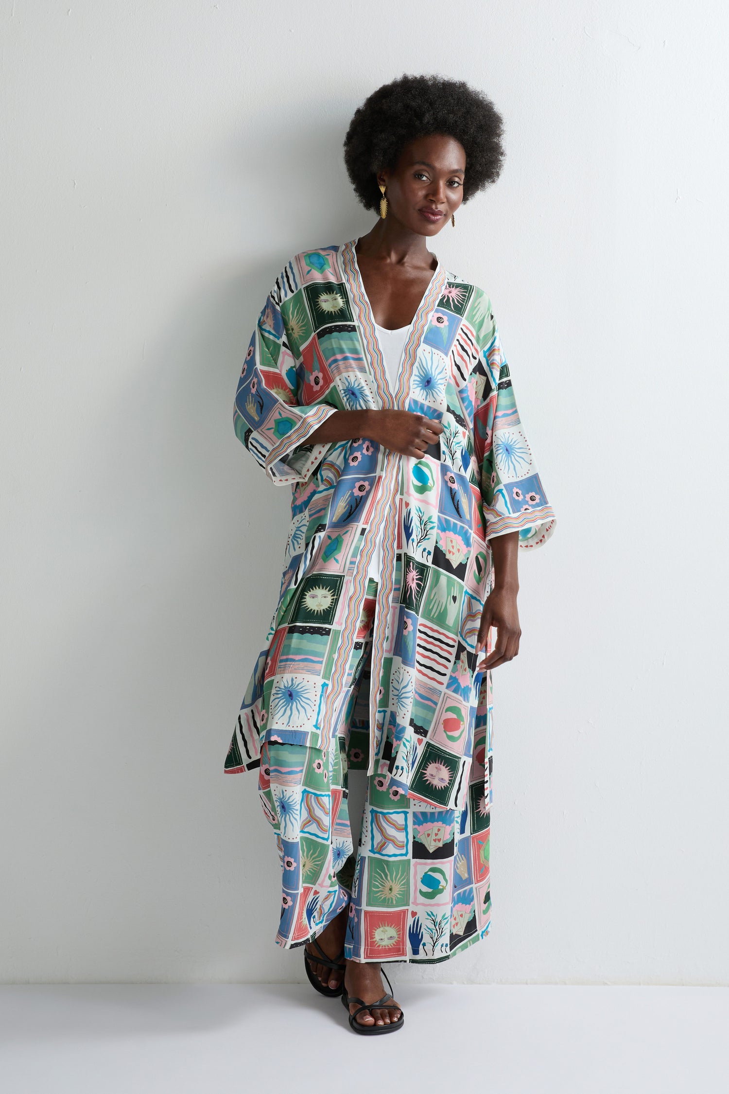 A woman stands by a white wall in the Mystical Collage Kimono, a long, loose piece with wide sleeves and black sandals, capturing free-spirited summer style.