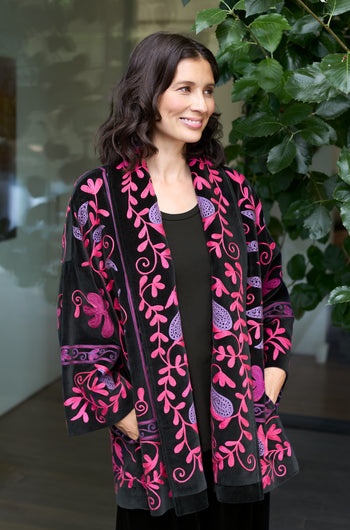 A woman stands indoors near a leafy plant, wearing a black top and the Winter Embroidered Jacket, which features striking bright pink and purple floral patterns.
