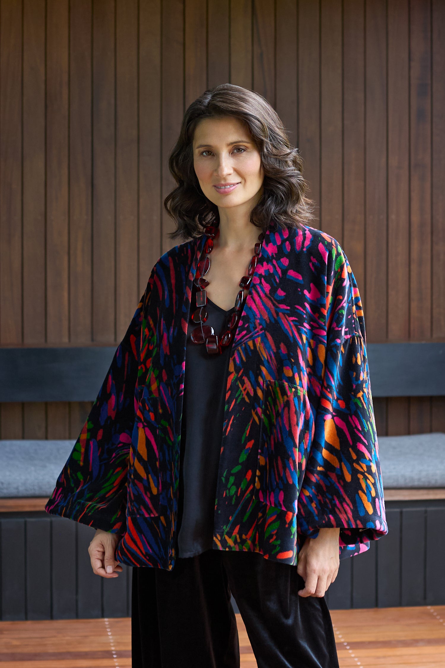 A woman with shoulder-length dark hair wears the Skyfire Velvet Velvet Jacket over a black top and necklace, standing indoors against a wooden wall—an inspiring pick for elevated evening looks.